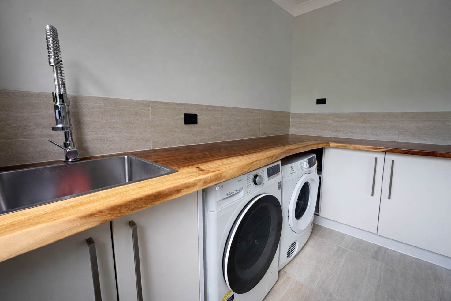 Laundry room with a stainless steel sink, wooden countertop, and a washing machine and dryer beneath the counter, with beige tiled walls and white cabinets.