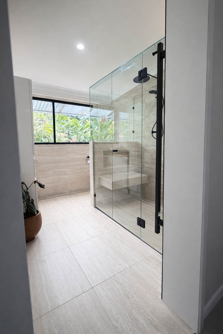 Modern walk-in shower with glass doors, beige tiles, bench, and a window showing greenery outside.