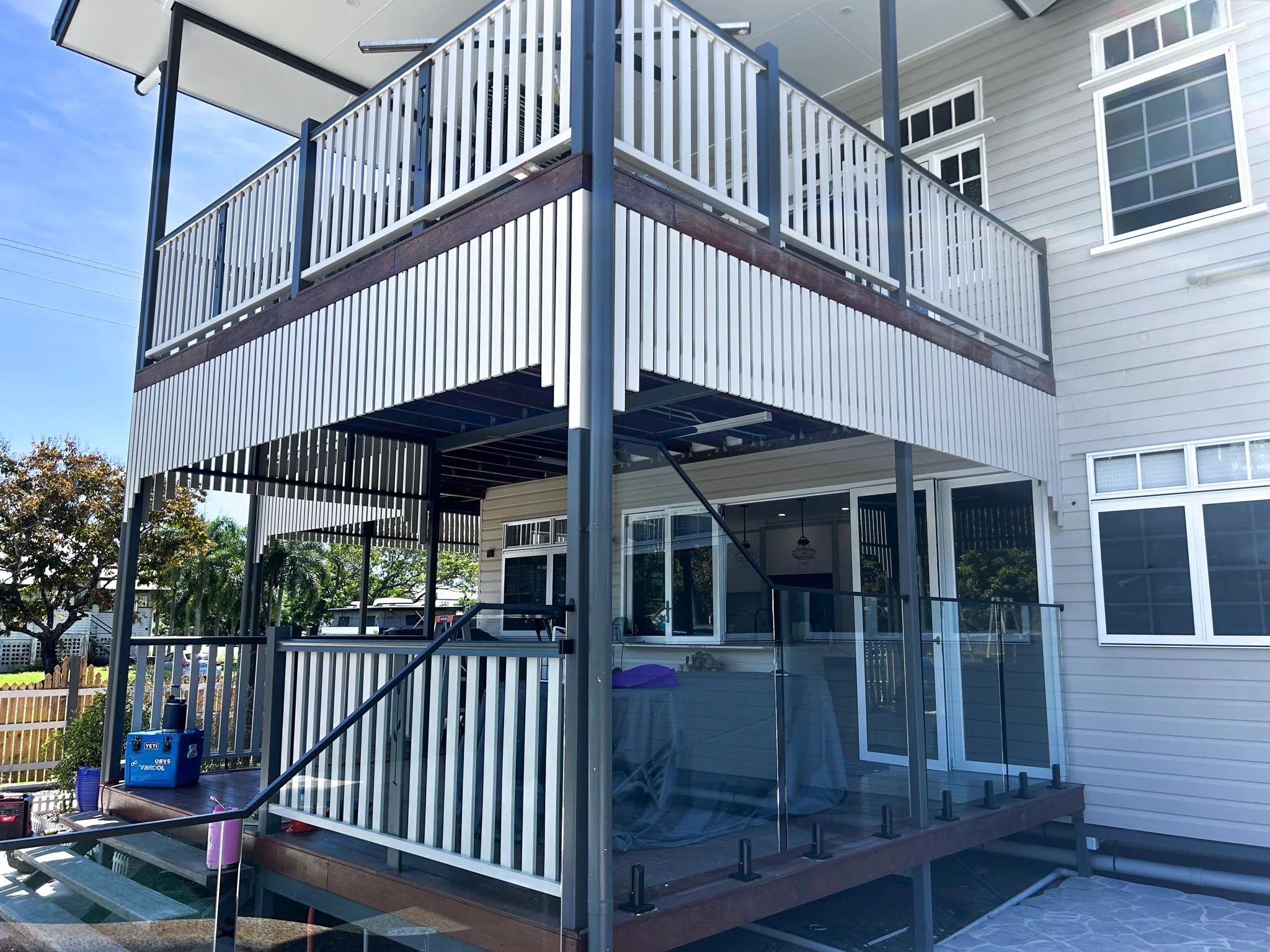 A two-story house with a balcony and staircase with glass railing, under construction or renovation, with tools and materials visible around and trees outside.
