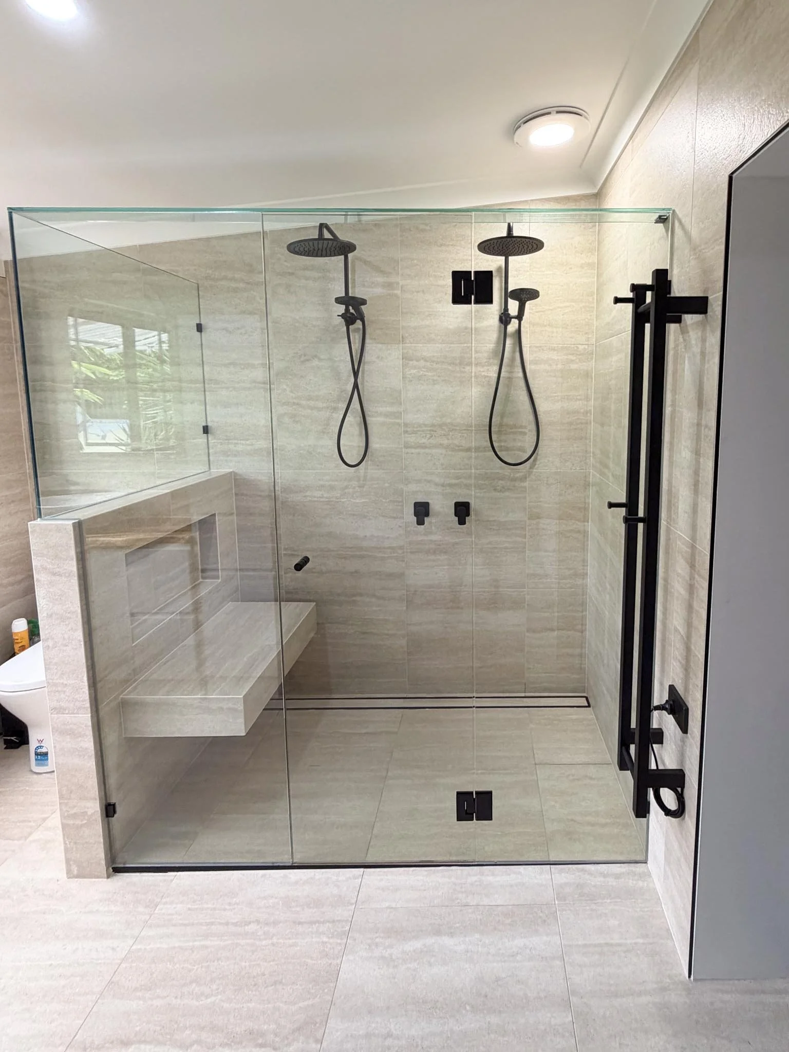 Modern glass shower with dual shower heads, black fixtures, and a built-in bench, beige tiled walls, and a gray cabinet outside the shower.