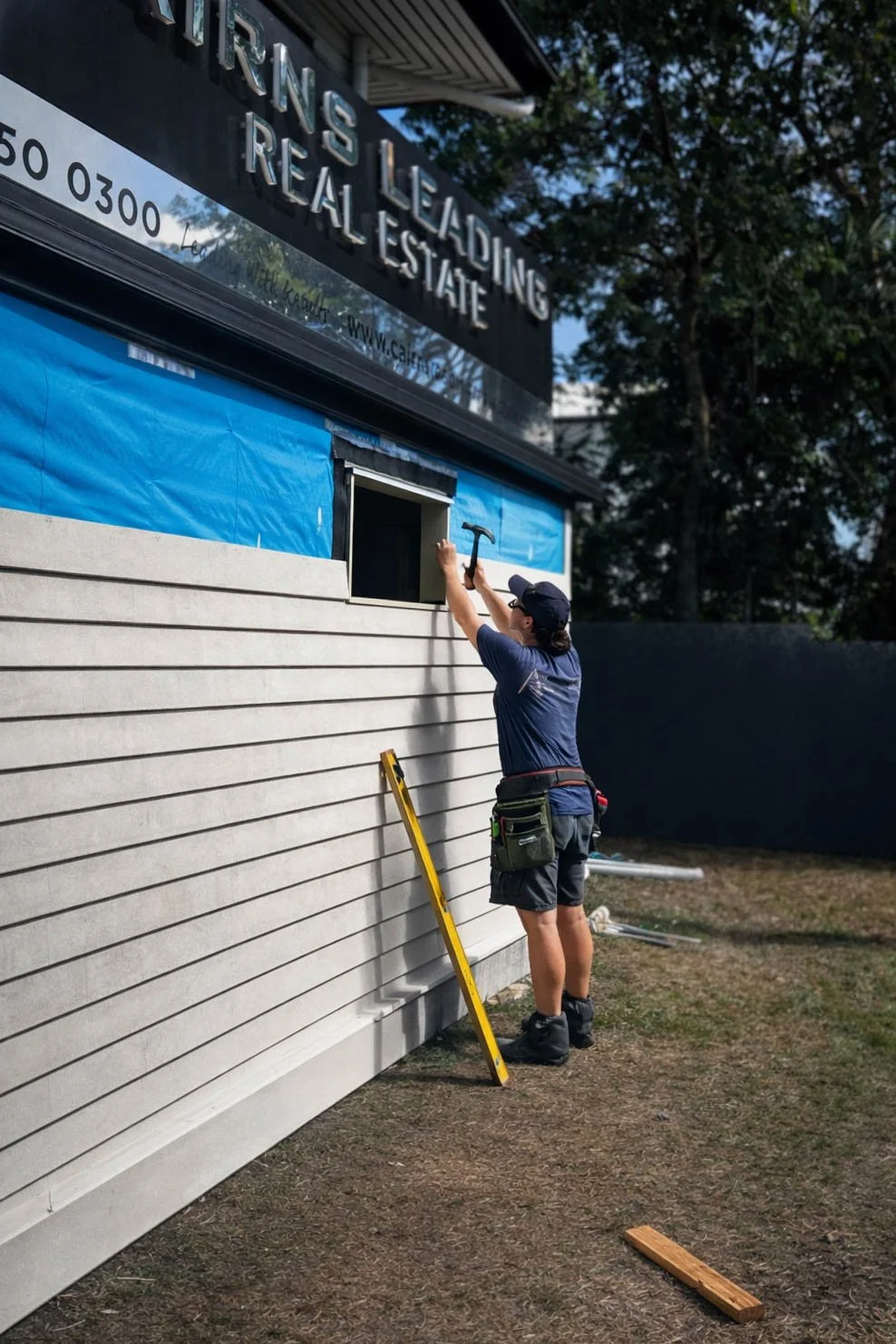 A worker from Birchwood Building Co installing siding on a building exterior, standing on the ground, using a hammer, with a ladder beside her, during daytime in Cairns.