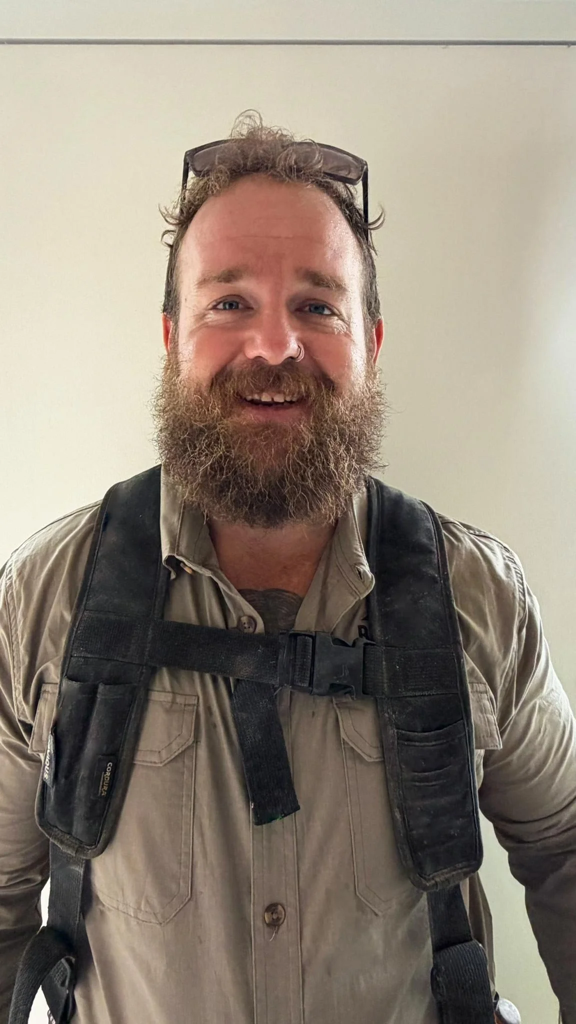 A man with curly hair and a full beard, wearing glasses on his head, a beige button-up shirt, and a black backpack, smiling in front of a plain light-colored wall.