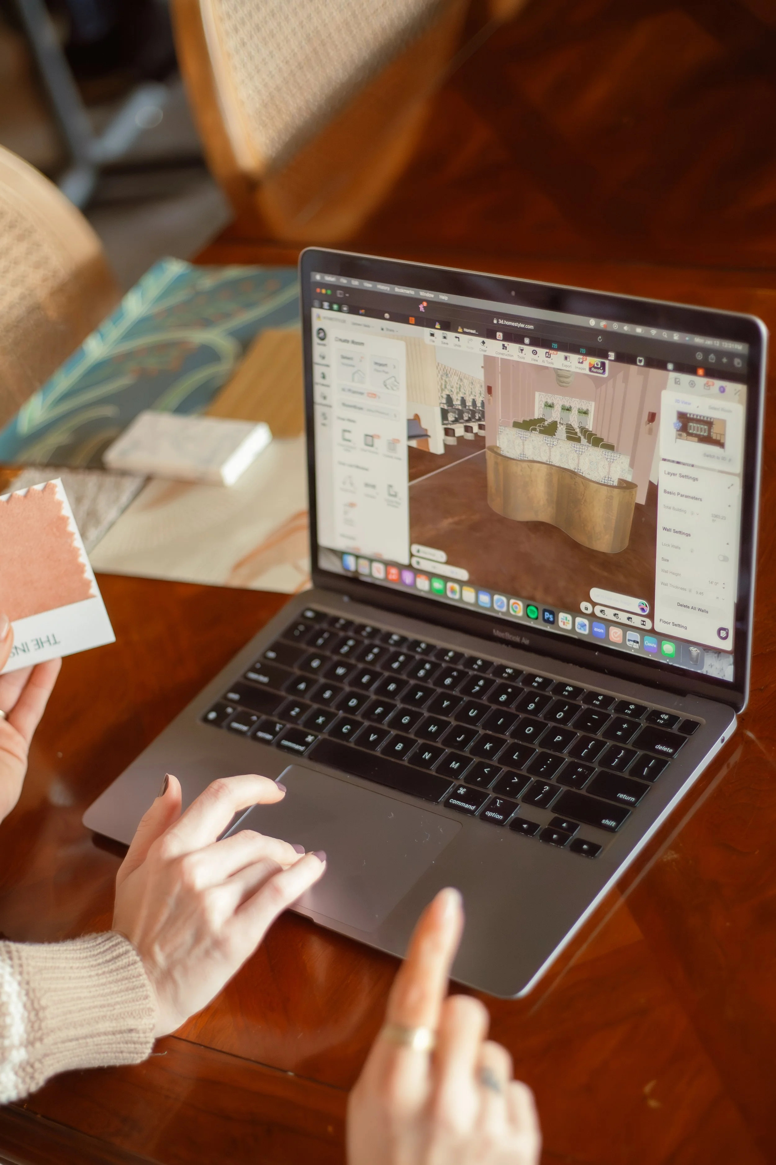 Laptop displaying 3D interior design of a reception area on a wooden table with fabric samples and a material sample card nearby.