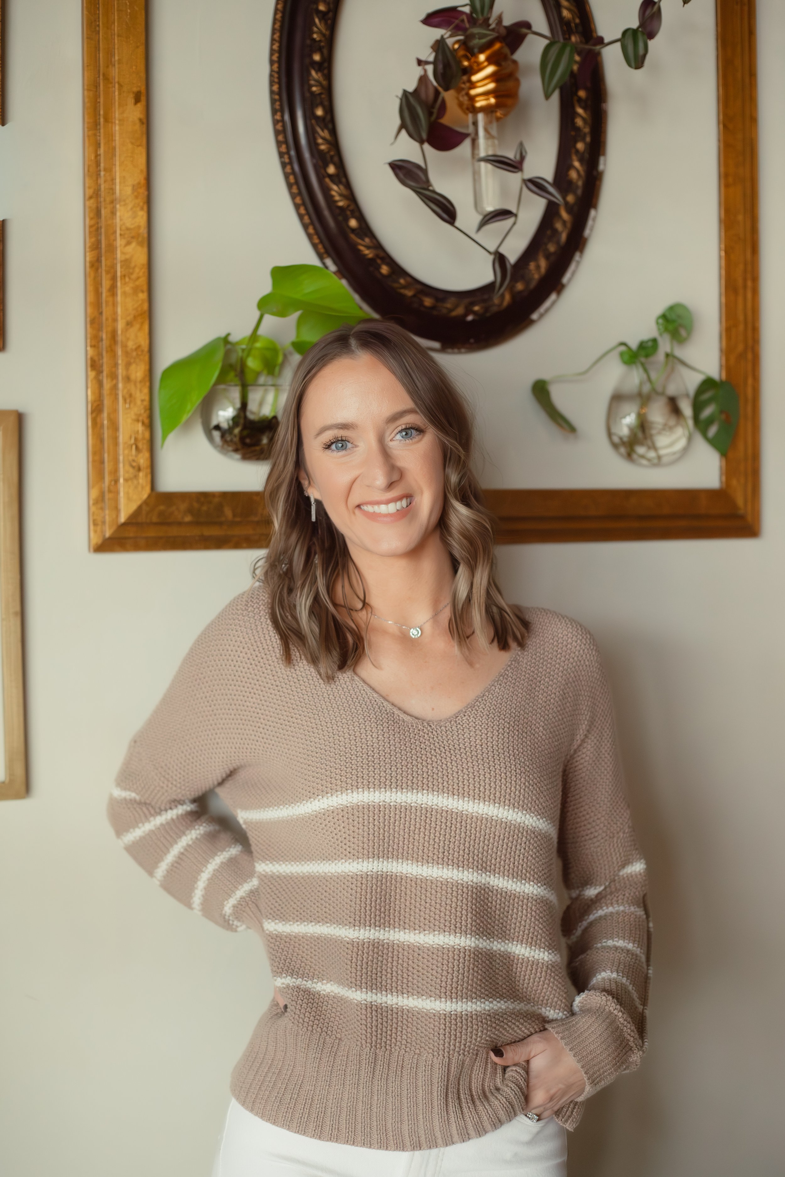 A woman with shoulder-length wavy brown hair and blue eyes, smiling and wearing a beige striped sweater and jewelry, standing against a wall with framed art and hanging plants.