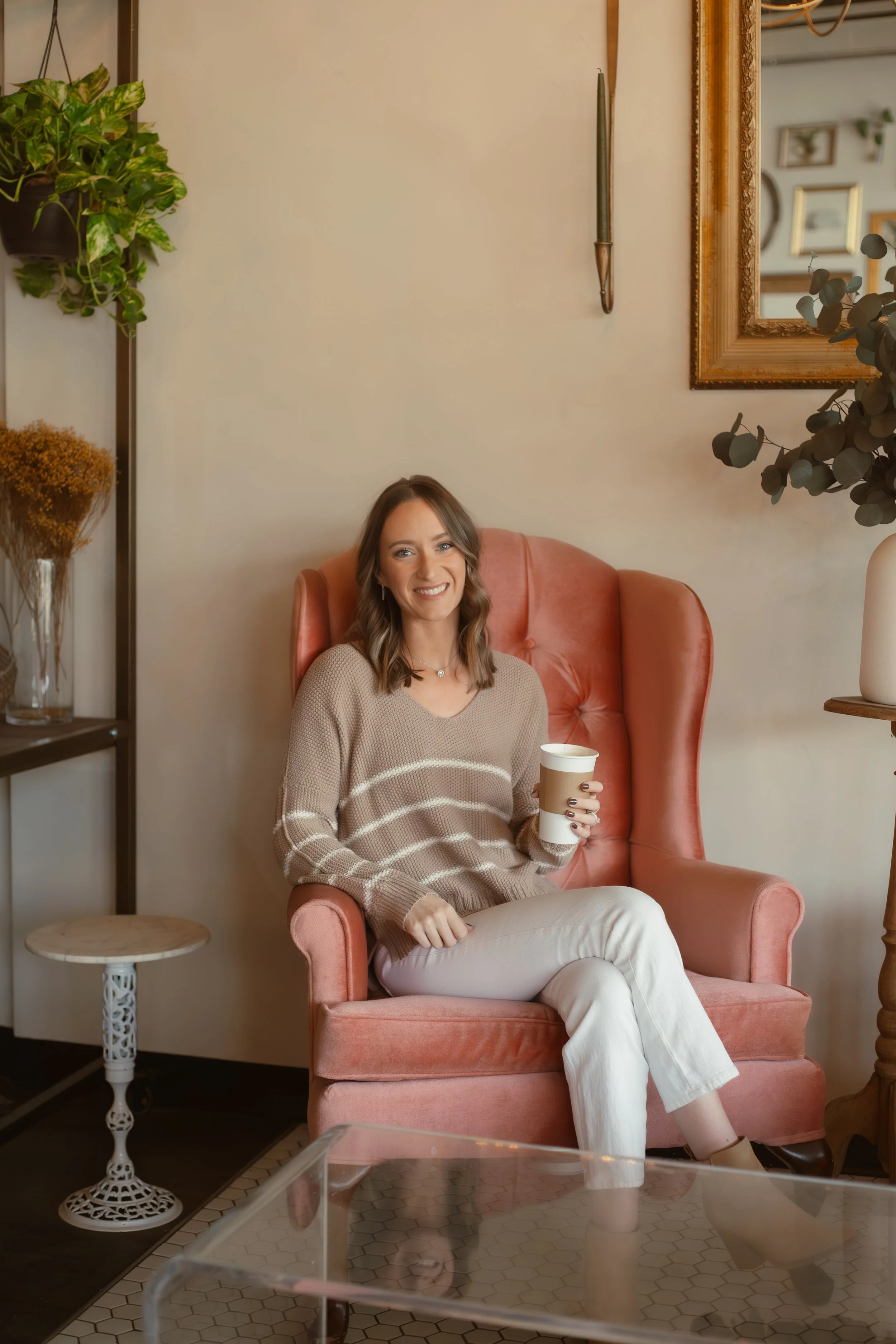A woman sitting in a pink armchair holding a coffee cup inside a cozy cafe with plants and framed art on the wall.