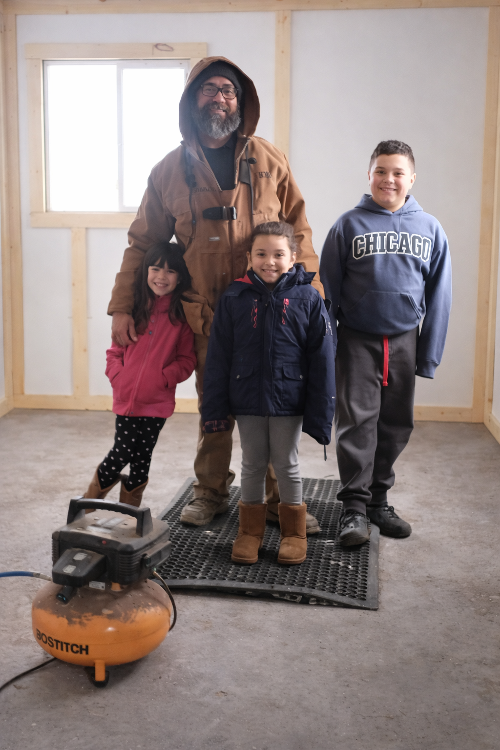 Man with three children inside a building under construction, with unfinished walls and a window in the background. The man is wearing a brown jacket, hoodie, glasses, and has a beard. The children are dressed in winter clothing, smiling, with an air compressor in the foreground.