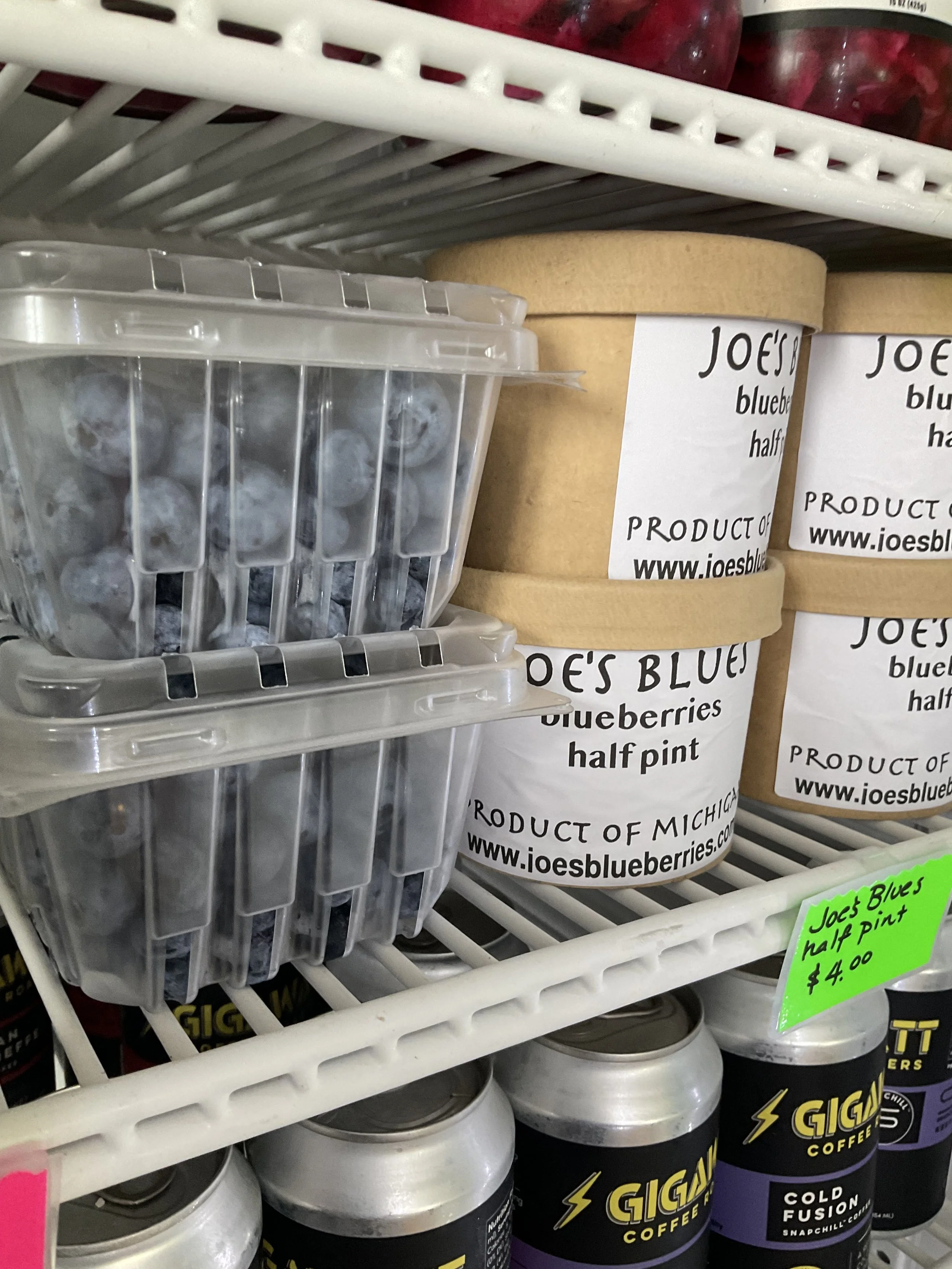 Frozen blueberries in clear plastic containers, labeled as Joe's Blueberries half pint, on a store shelf. There are also coffee cans labeled GIGANTIC Coffee on the bottom shelf.