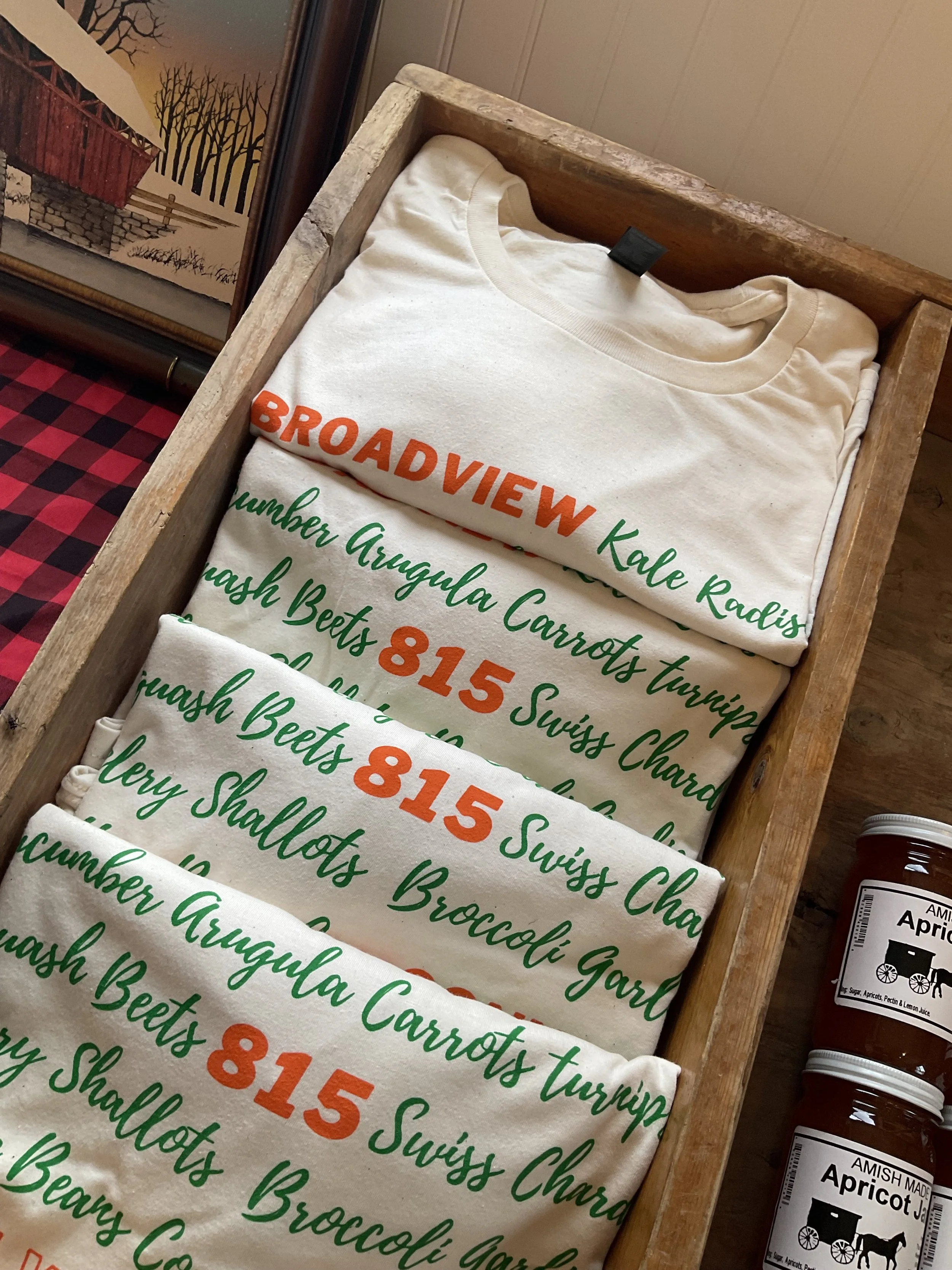 Folded cream-colored t-shirts with green and orange text in a wooden box, with jars of apricot jam nearby. The text on the shirts reads 'BROADVIEW Kale Radish,' '815 Swiss Chard,' and 'Brussels Sprouts'.