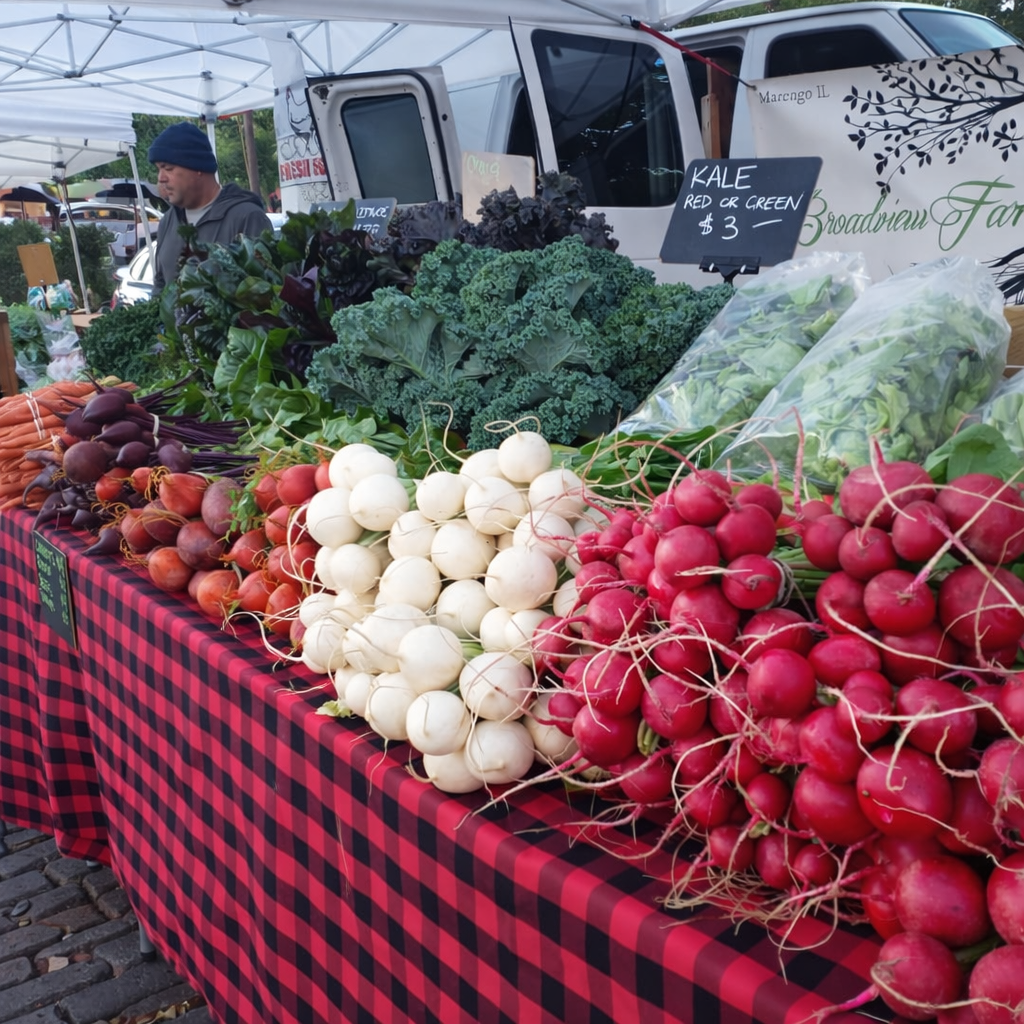 Fresh vegetables displayed at an outdoor farmers market stall, including carrots, beets, turnips, and radishes, with some leafy greens in plastic bags. A man is seen in the background browsing the produce.