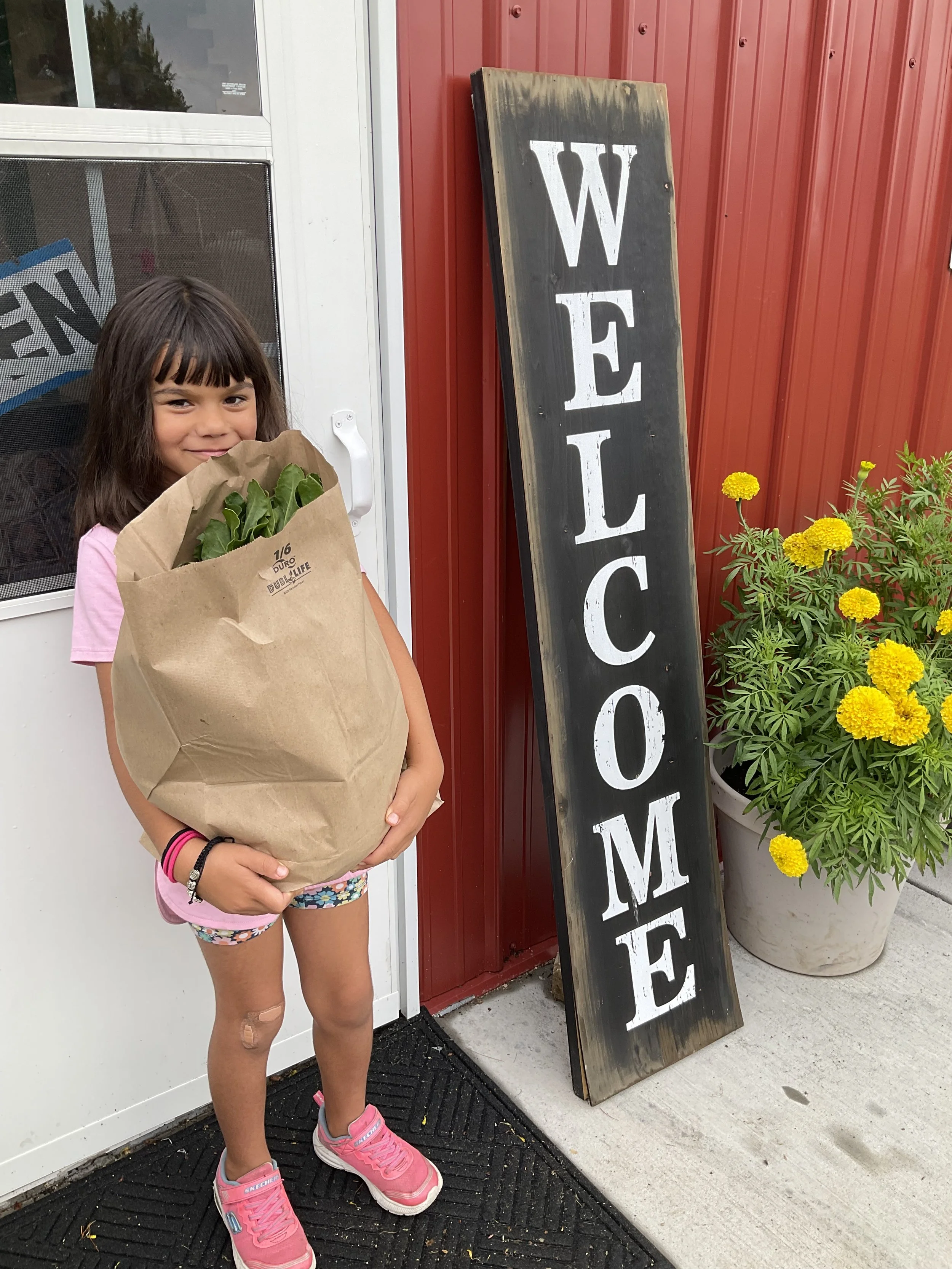 A young girl with a band-aid on her leg, holding a paper bag with greens inside, standing in front of a store with a welcome sign, near a potted yellow flower.
