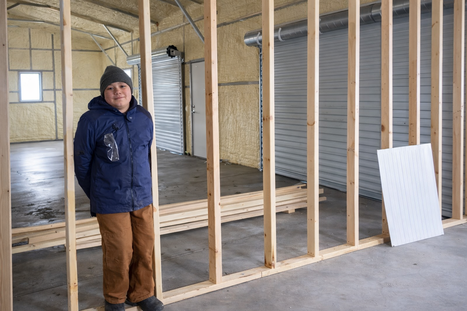 A boy wearing a gray beanie and a blue jacket stands inside a partially constructed room with wooden framing, a concrete floor, and insulation on the walls.