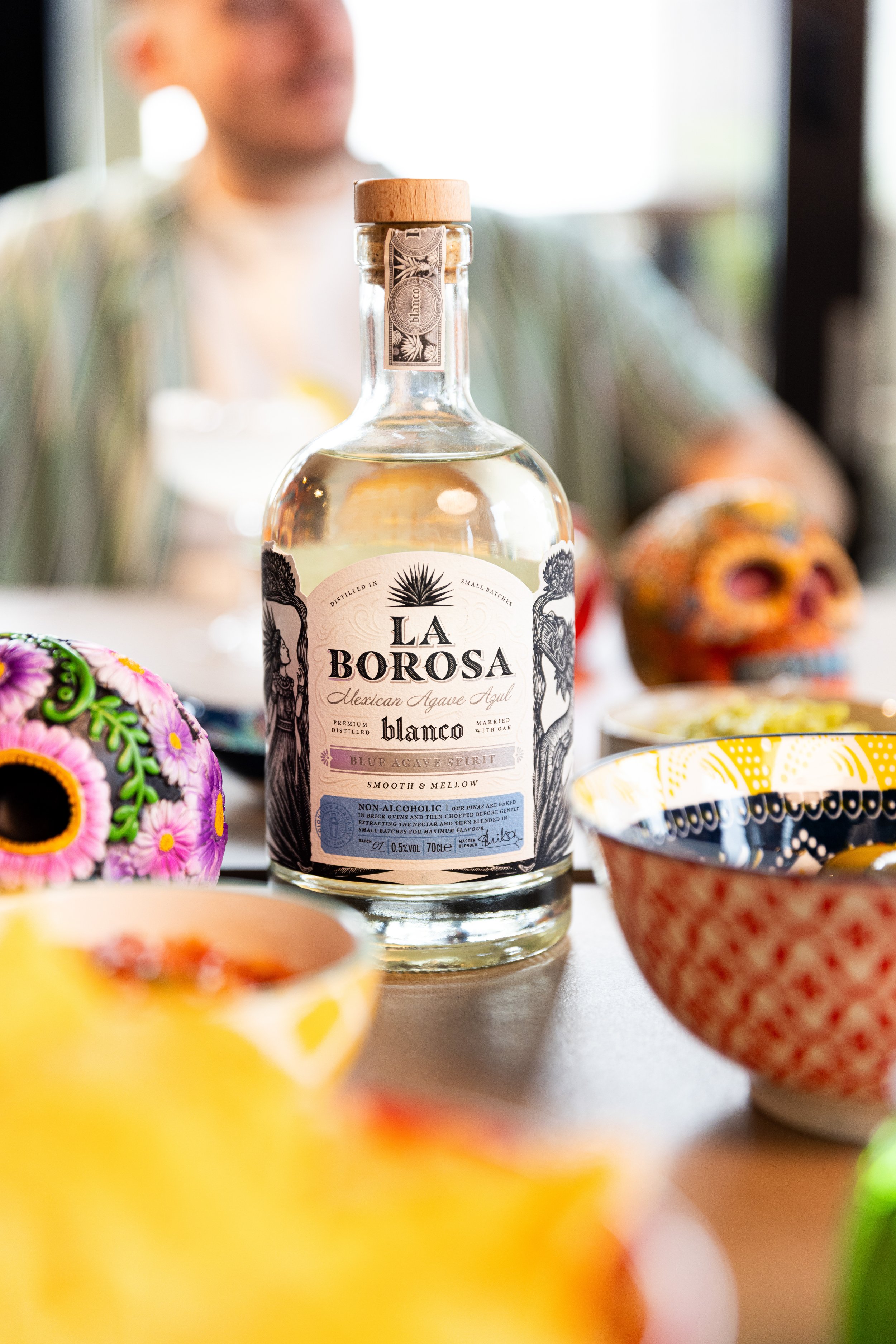 A bottle of La Borosa Blue Agave Spirit with Mexican-themed artwork on the label, surrounded by colorful decorative skulls and bowls, with a blurred person in the background.