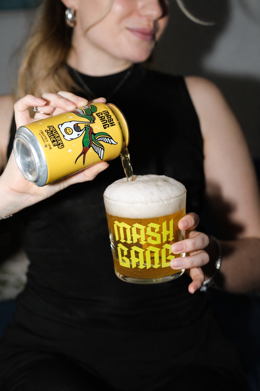 A woman in black clothing is pouring a beer from a can into a glass mug with foam on top. The can has colorful artwork featuring jaguars and green birds, with the label "Electric Boogie." The glass mug has the words "Mash Bash" written on it in yellow.