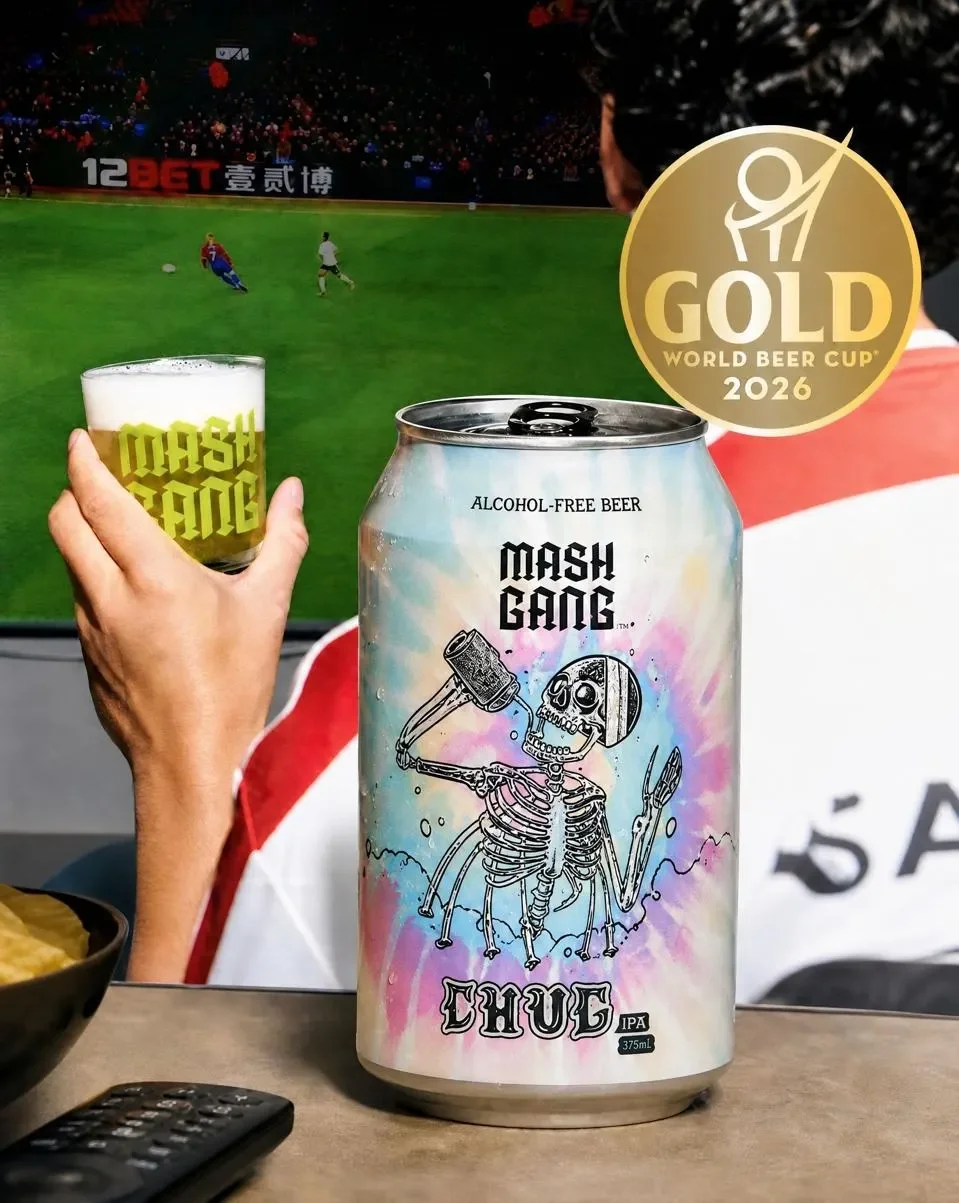 A can of Mash Gang alcohol-free beer in front of a TV showing a soccer game. A person is holding a glass of beer labeled 'WOSH SAID.' There is a gold medal graphic indicating a 2026 World Beer Cup gold award. The can design features a skeleton holding a mallet and knife with a colorful background and the words Morph Gang and Chufe IPA.