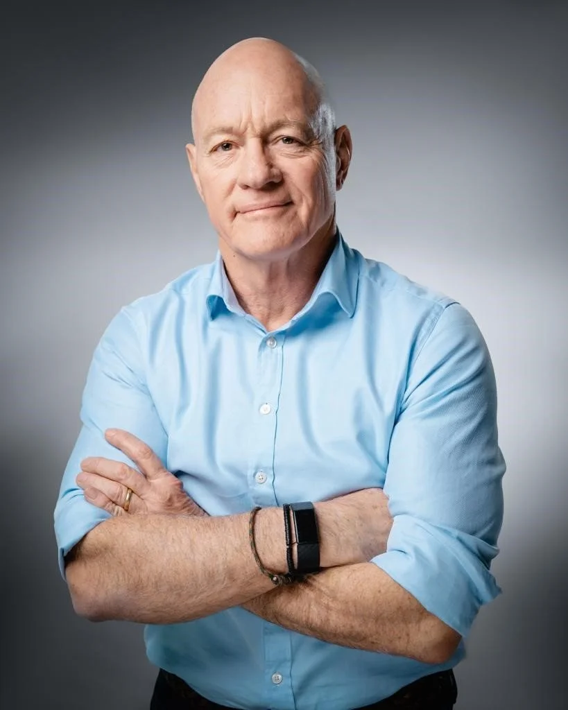 A middle-aged man with a bald head wearing a light blue button-up shirt, standing with arms crossed against a gray gradient background.