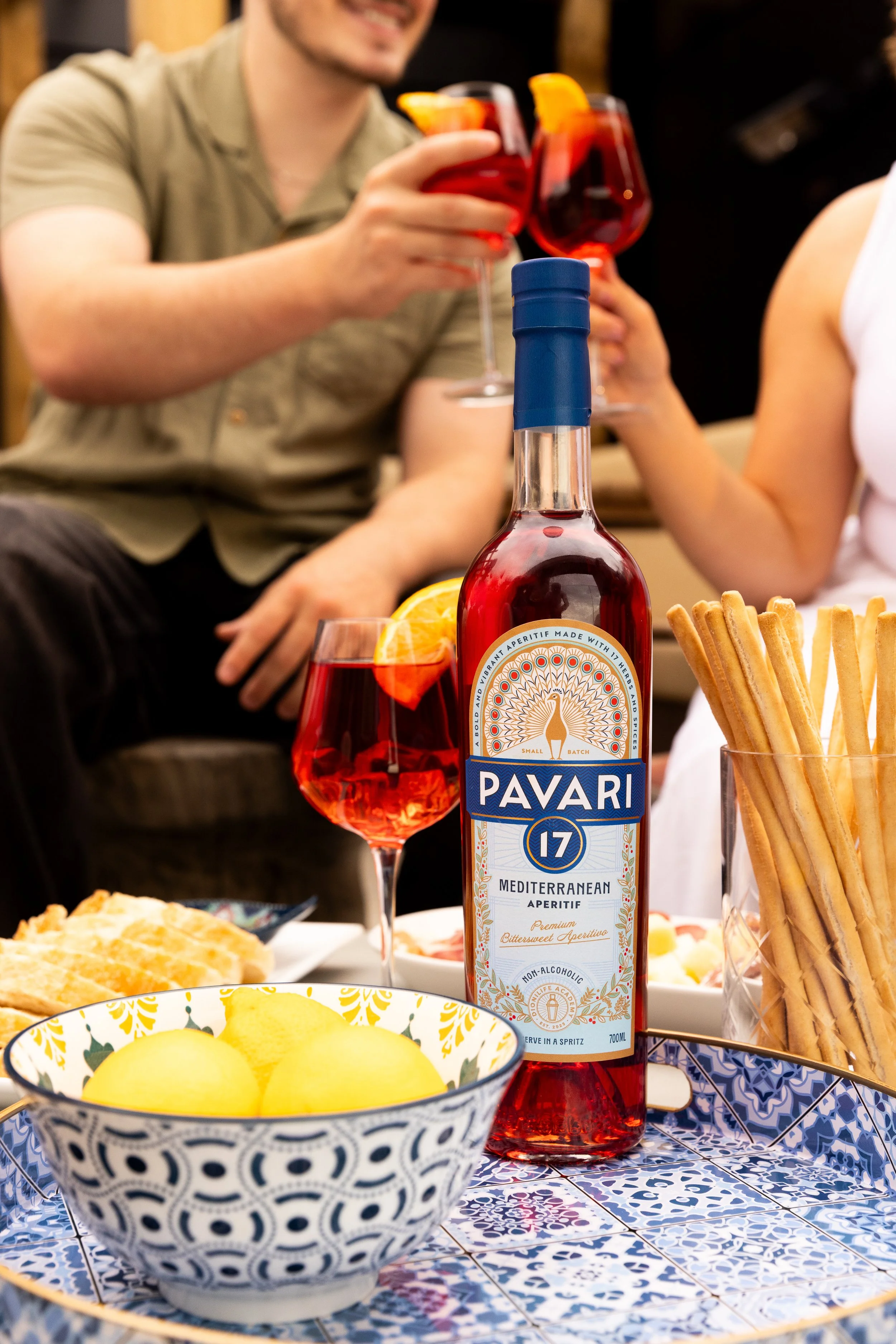 People enjoying a meal with a bottle of Pavari Mediterranean aperitif and various snacks, including a bowl of yellow melon balls and breadsticks, on a blue and white patterned table.