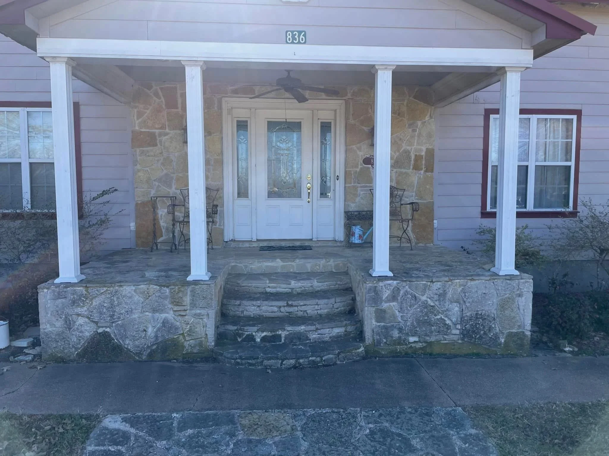 Front porch of a house with stone steps, white columns, a white front door with glass panels, two chairs, a small table, and a ceiling fan.