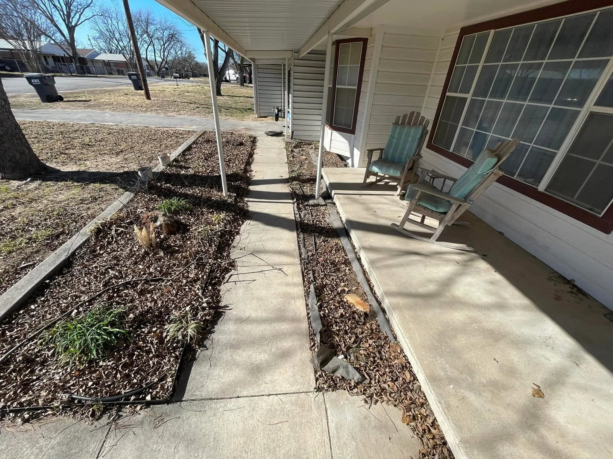 Empty front porch with two wooden chairs with green cushions, a small table, shadow of a tree, and a flower bed with dried leaves and some plants, in a neighborhood with houses and leafless trees.