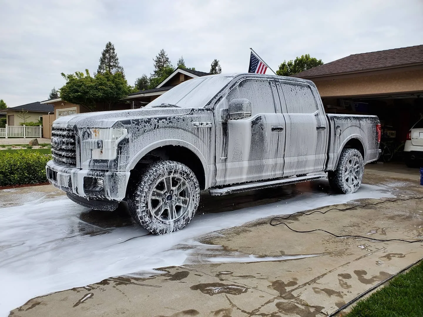 A large pickup truck covered in soap suds during a wash, parked outside a house with an American flag on a pole, amid a cloudy sky.