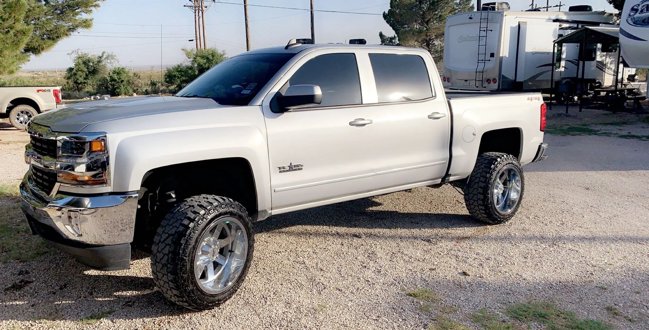 Silver pickup truck with large tires parked on gravel at a campsite with RVs and trees in the background.