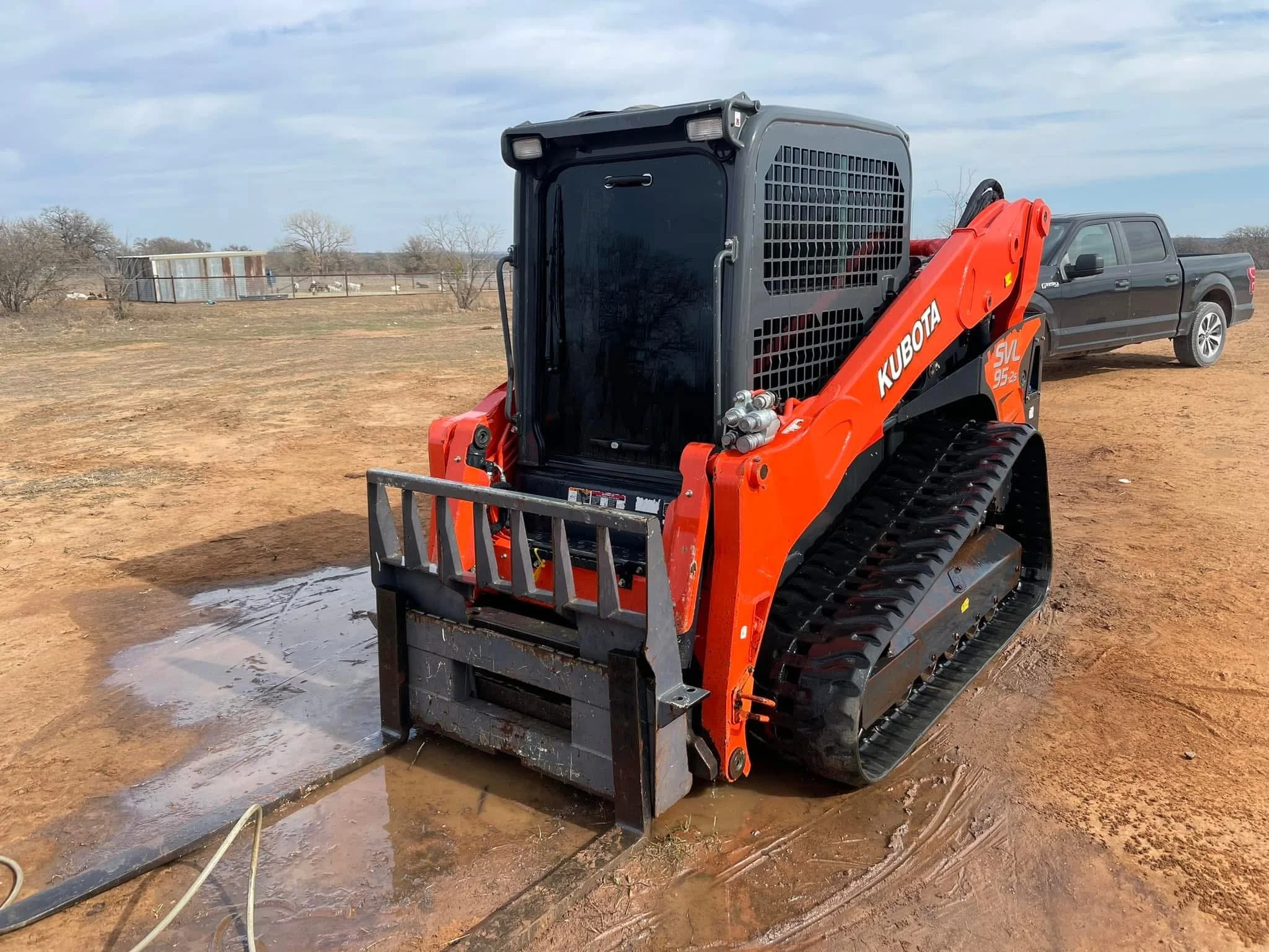 A Kubota SVL 95-2 compact track loader with an orange body and black tracks, parked on a muddy dirt ground, with a gray pickup truck in the background under a cloudy sky.