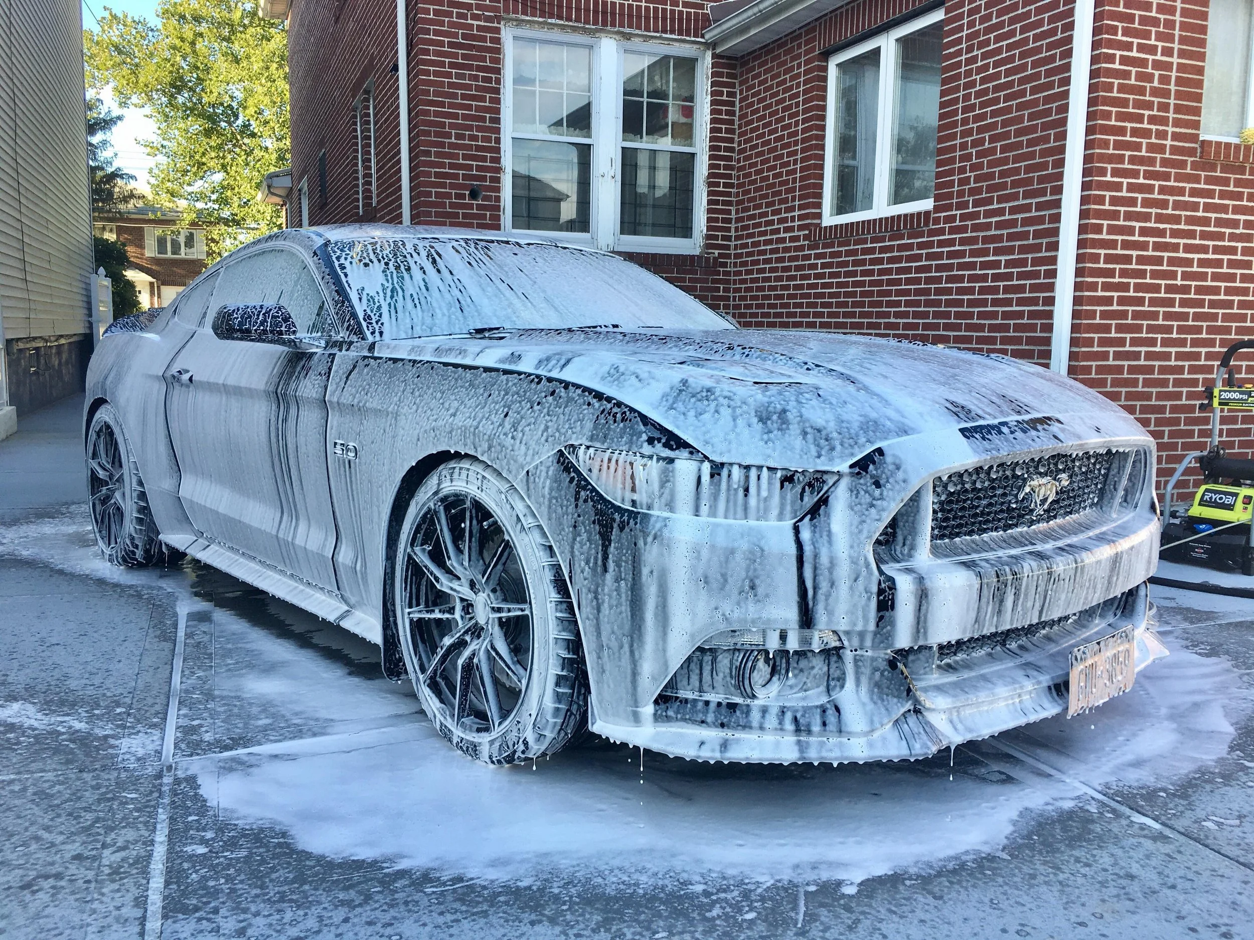 A black sports car with foam and soap suds on it during a car wash, parked on a concrete driveway in front of a brick house, with a pressure washer visible on the side.