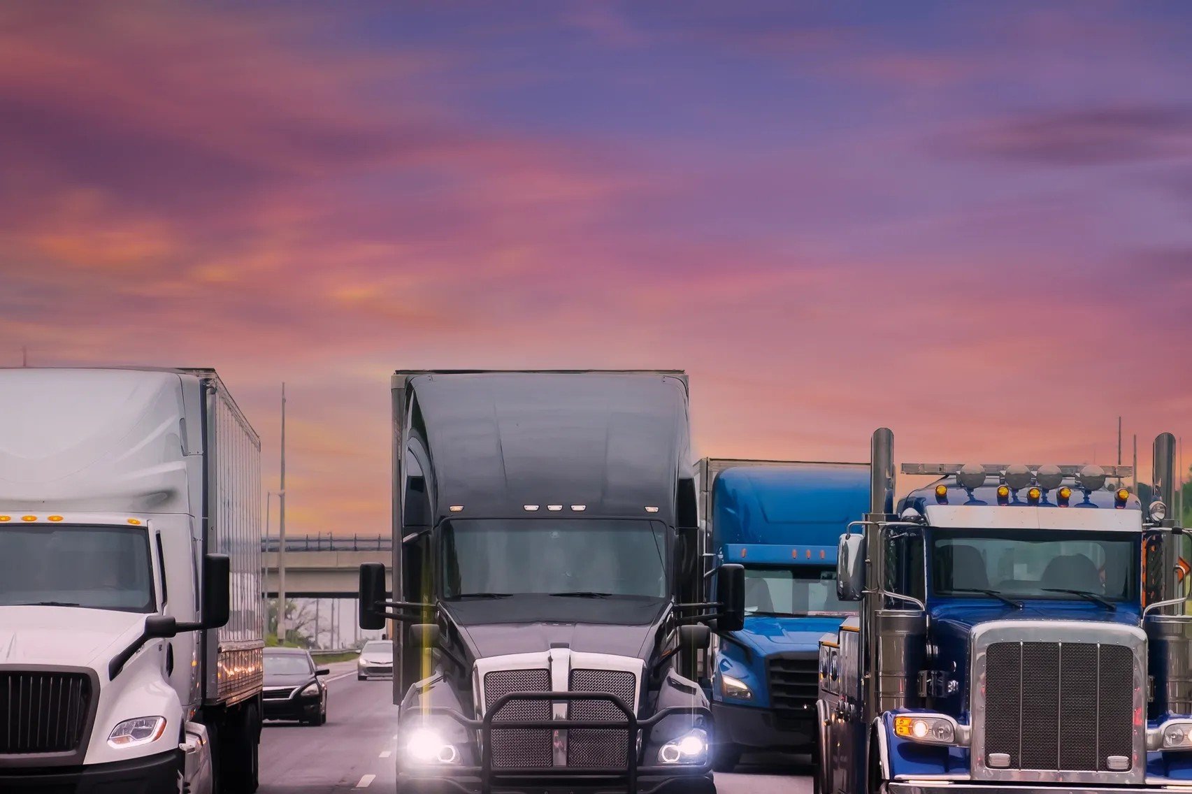 Row of trucks parked side by side on a highway during sunset with a purple and pink sky.