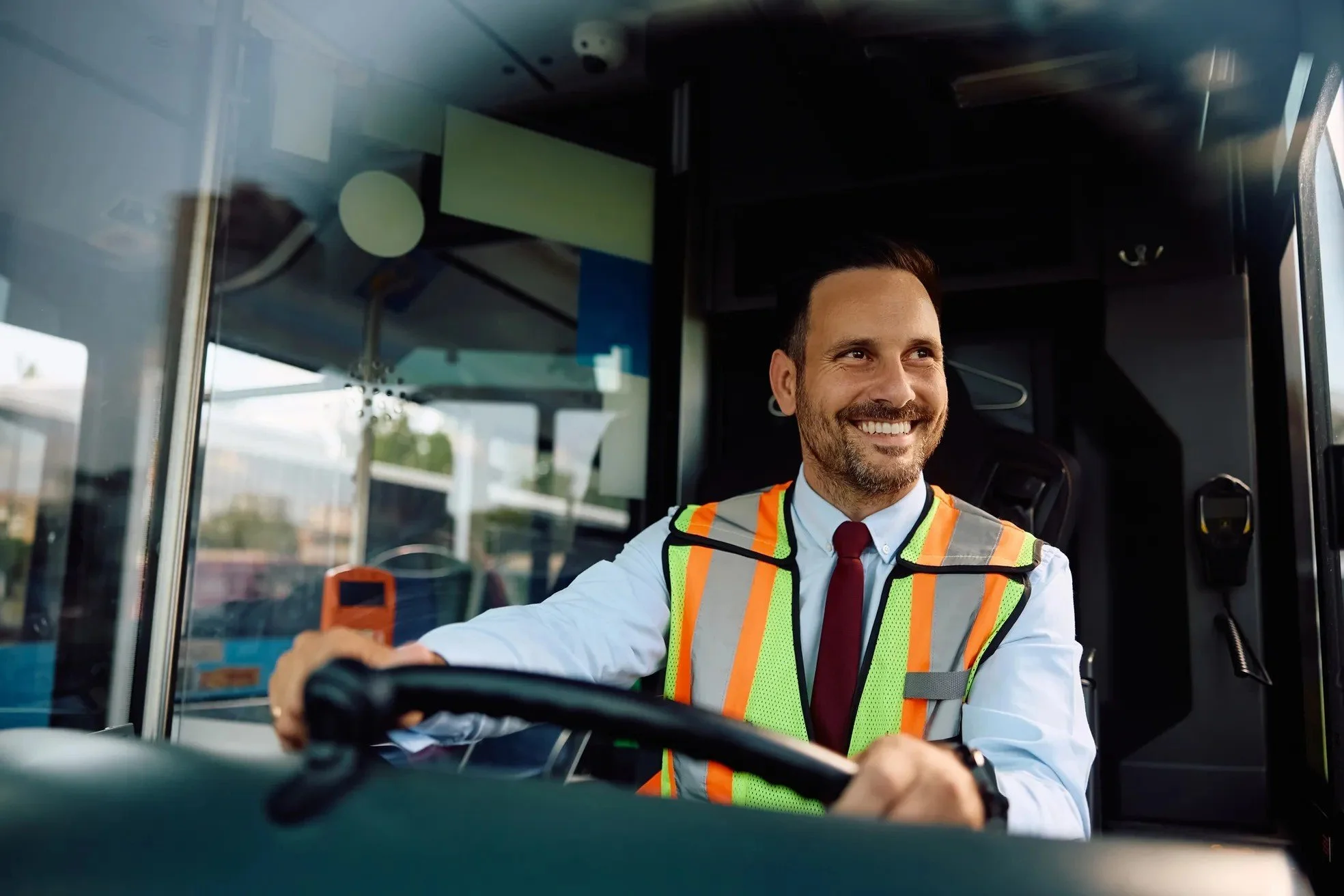 A smiling man in a safety vest driving a bus.