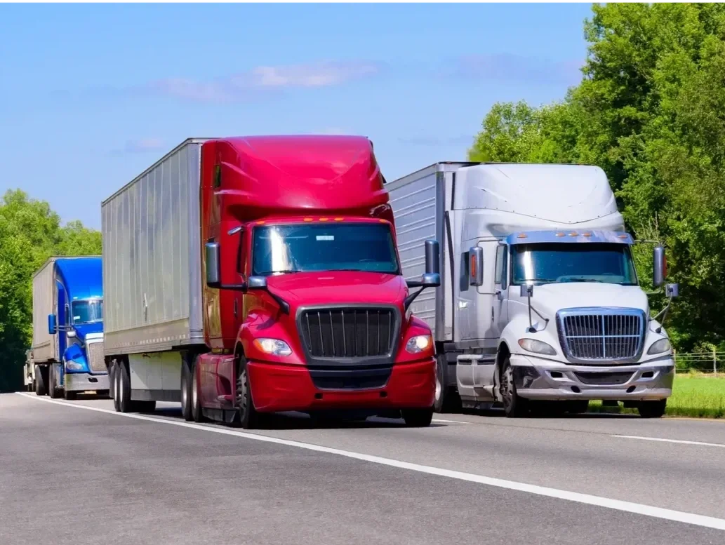 Three semi-trucks parked on the side of a two-lane road with green trees in the background, one in red, one in white, and one in blue.