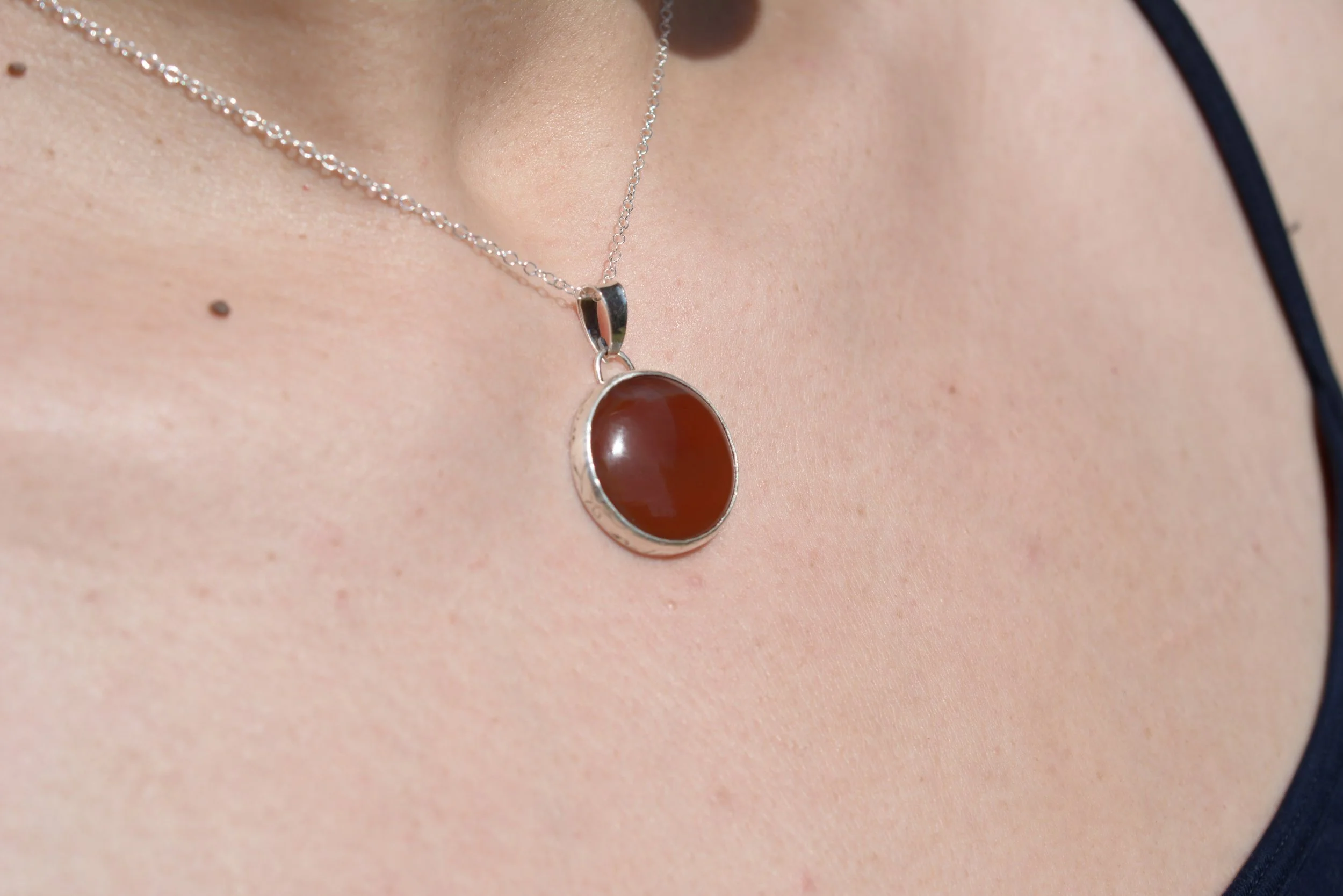 Close-up of a woman wearing a silver necklace with a large, oval, dark red carnelian pendant, and a black tank top with spaghetti straps.