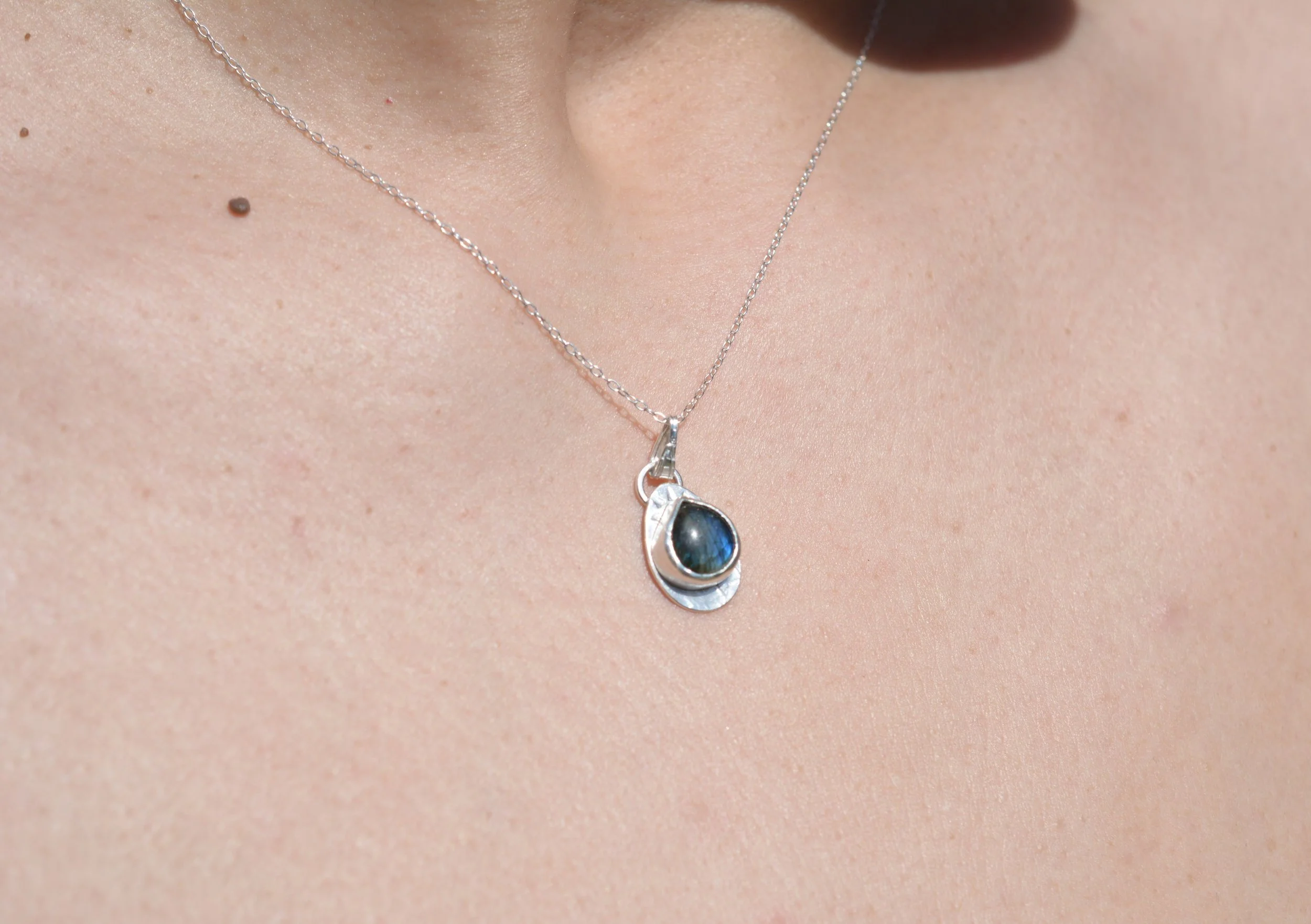Close-up of a woman's neck with a silver necklace featuring a teardrop-shaped dark labradorite gemstone pendant. set on a wide patterned silver bezel