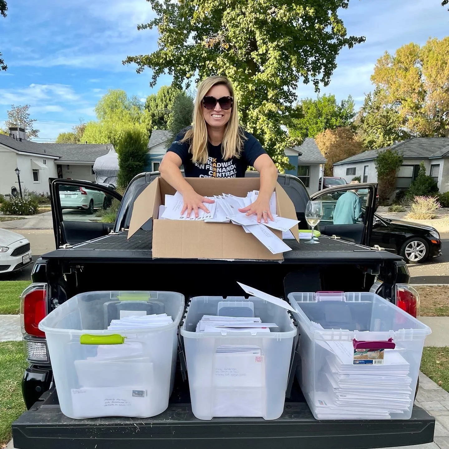 Woman with blonde hair and sunglasses on the back of a black pickup truck, with large plastic storage bins filled with Vote Forward letters ready to send.