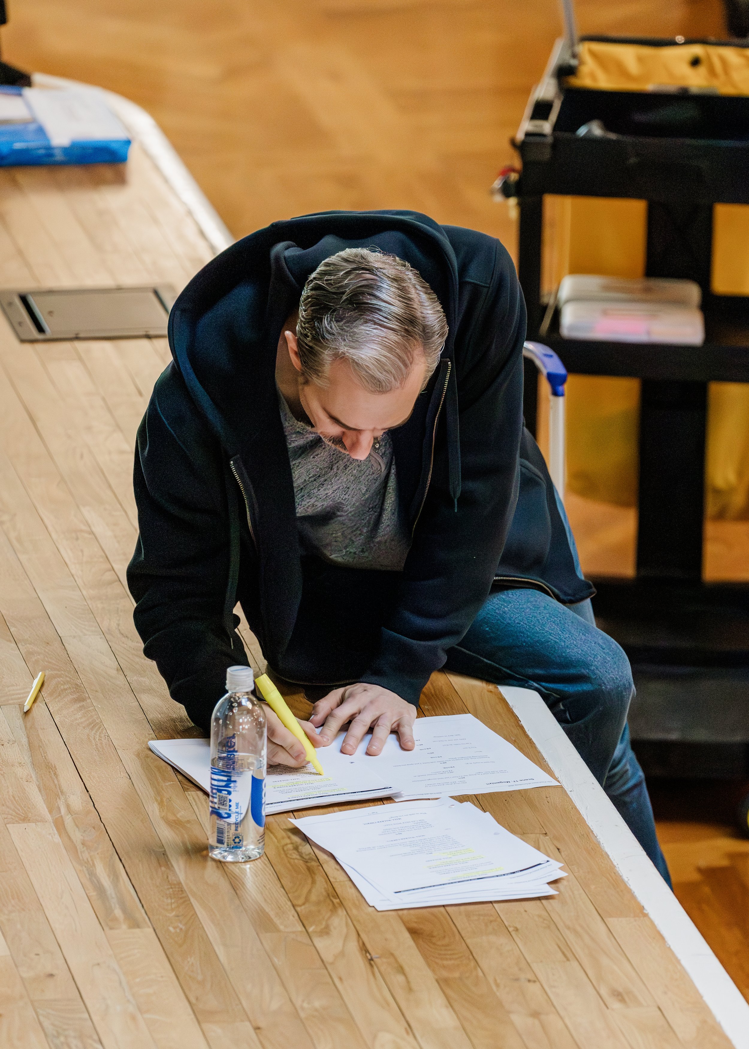 A man with gray hair, wearing a black hoodie and gray jeans, is kneeling on a wooden floor, writing on a sheet of paper with a yellow marker. There is a water bottle, some documents, and a yellow marker on the floor. Behind him is a rolling cart with books and a black and yellow suitcase.