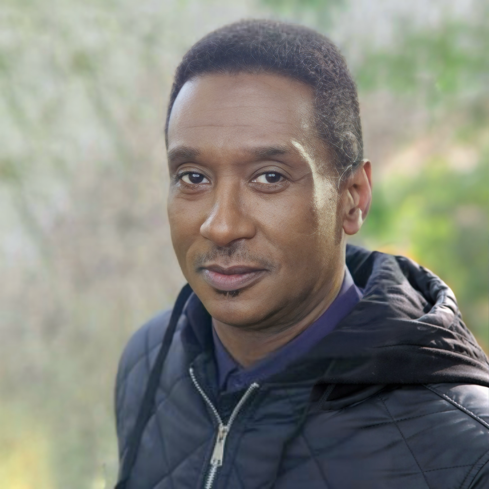 A man with short black hair, light brown skin, and a small goatee, wearing a navy blue jacket, standing outdoors with blurred green background.