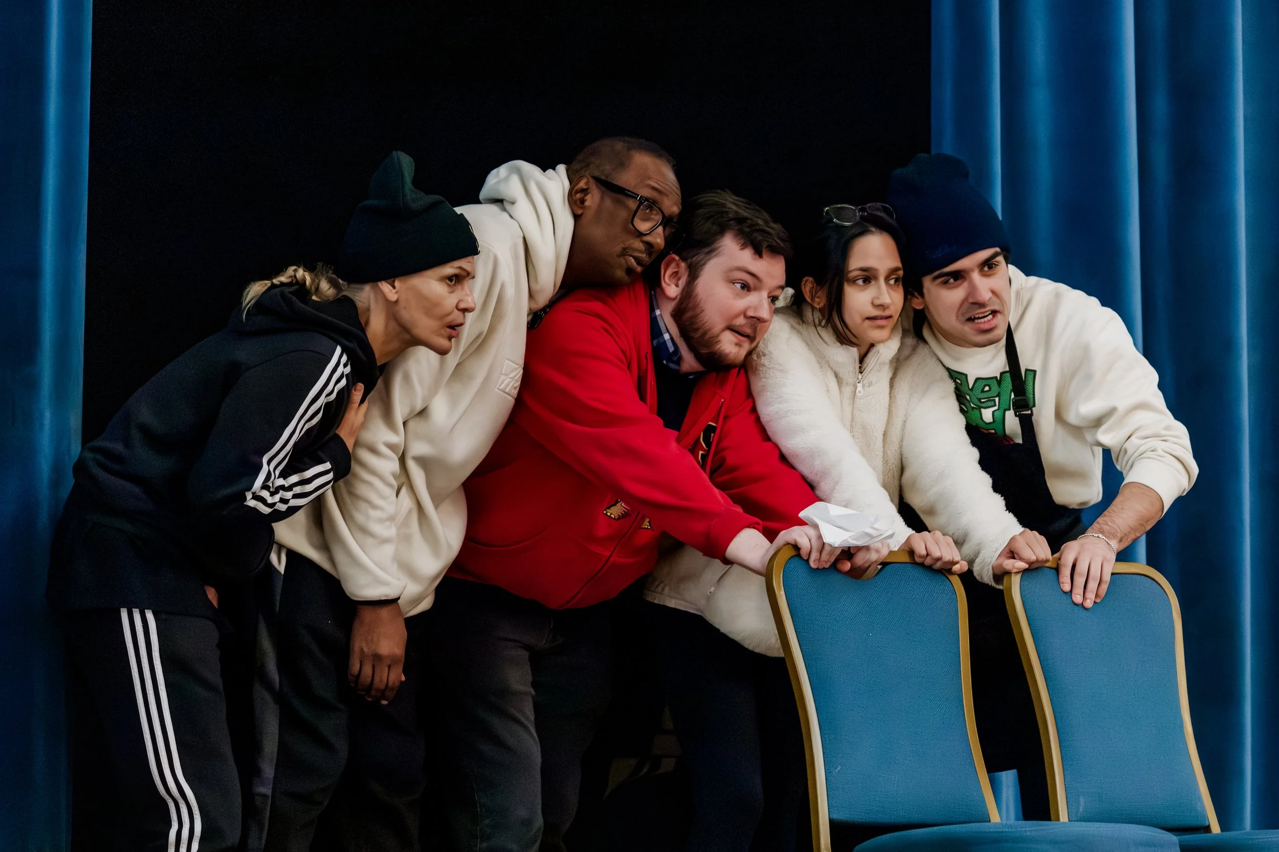 Five people huddled together on a stage, leaning forward and appearing surprised or intrigued, with blue chairs and curtain in the background.