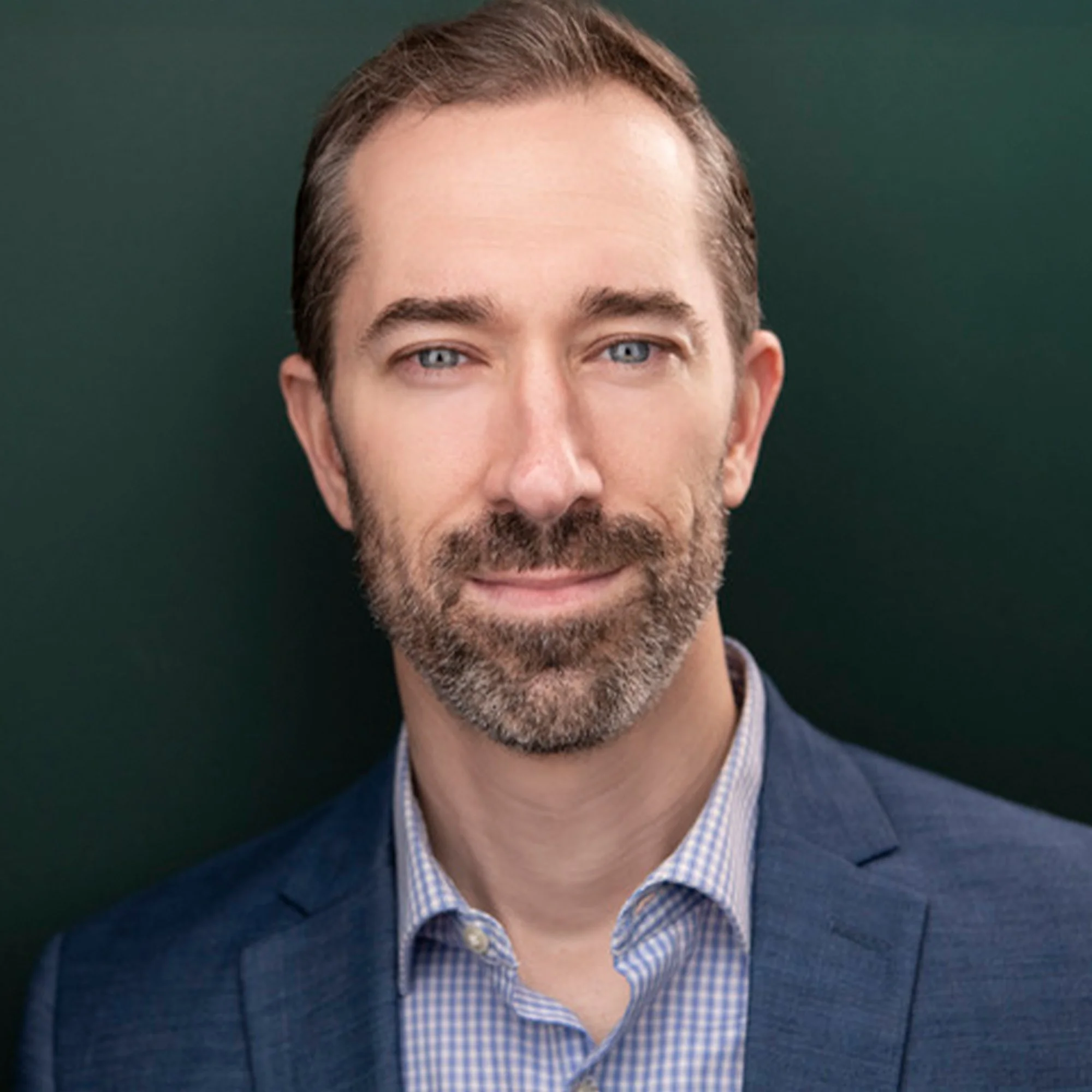 A portrait of a middle-aged man with blue eyes, short brown hair, and a beard, wearing a blue suit jacket and a light blue checkered shirt, against a dark green background.