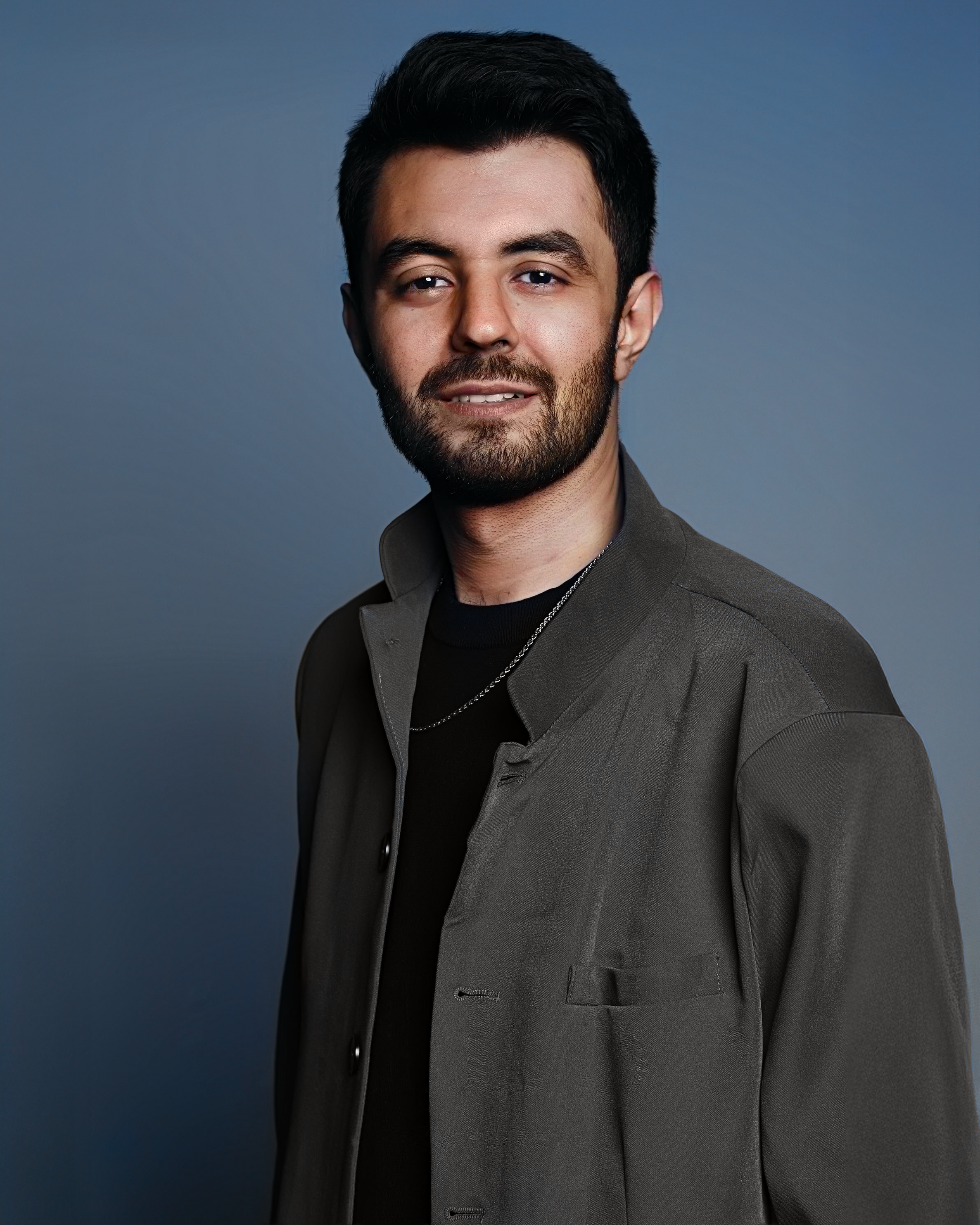 Portrait of a young man with short dark hair, beard, wearing a dark jacket over a black shirt, standing against a plain blue background.