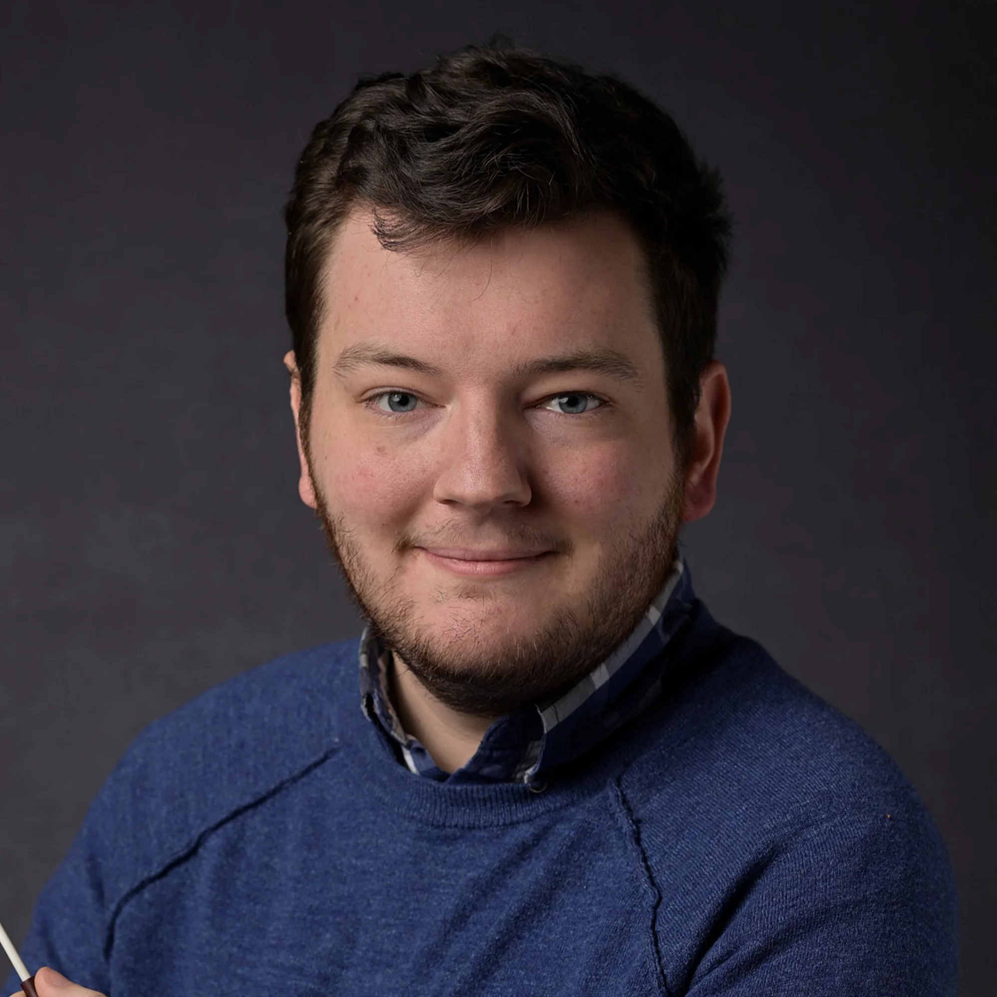Close-up portrait of a young man with brown hair and blue eyes, wearing a blue sweater over a checkered shirt, smiling softly against a dark background.