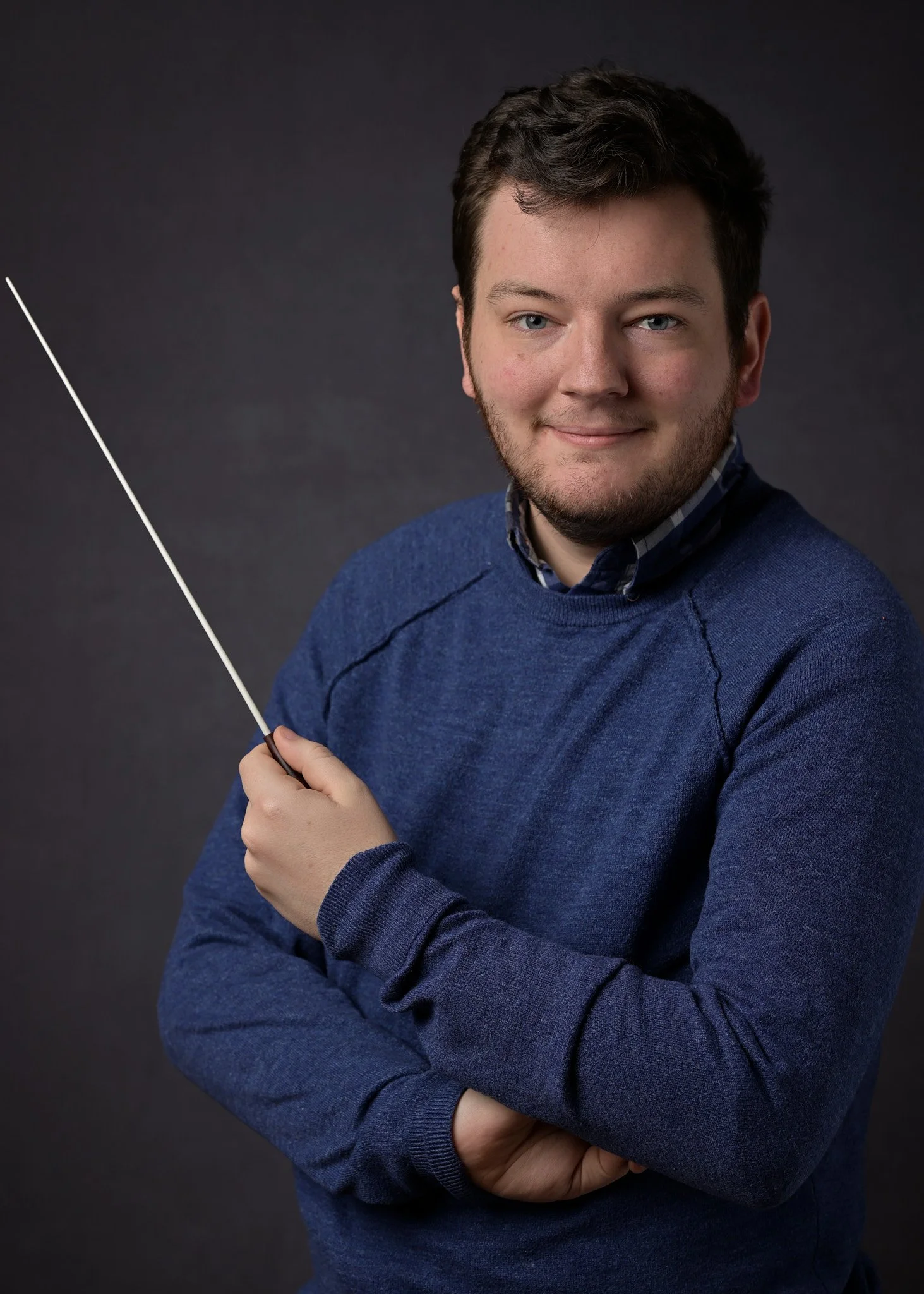 Young man with short dark hair and beard, wearing a blue sweater over a collared shirt, holding a thin pointer, standing against a dark grey background, looking at the camera and smiling.