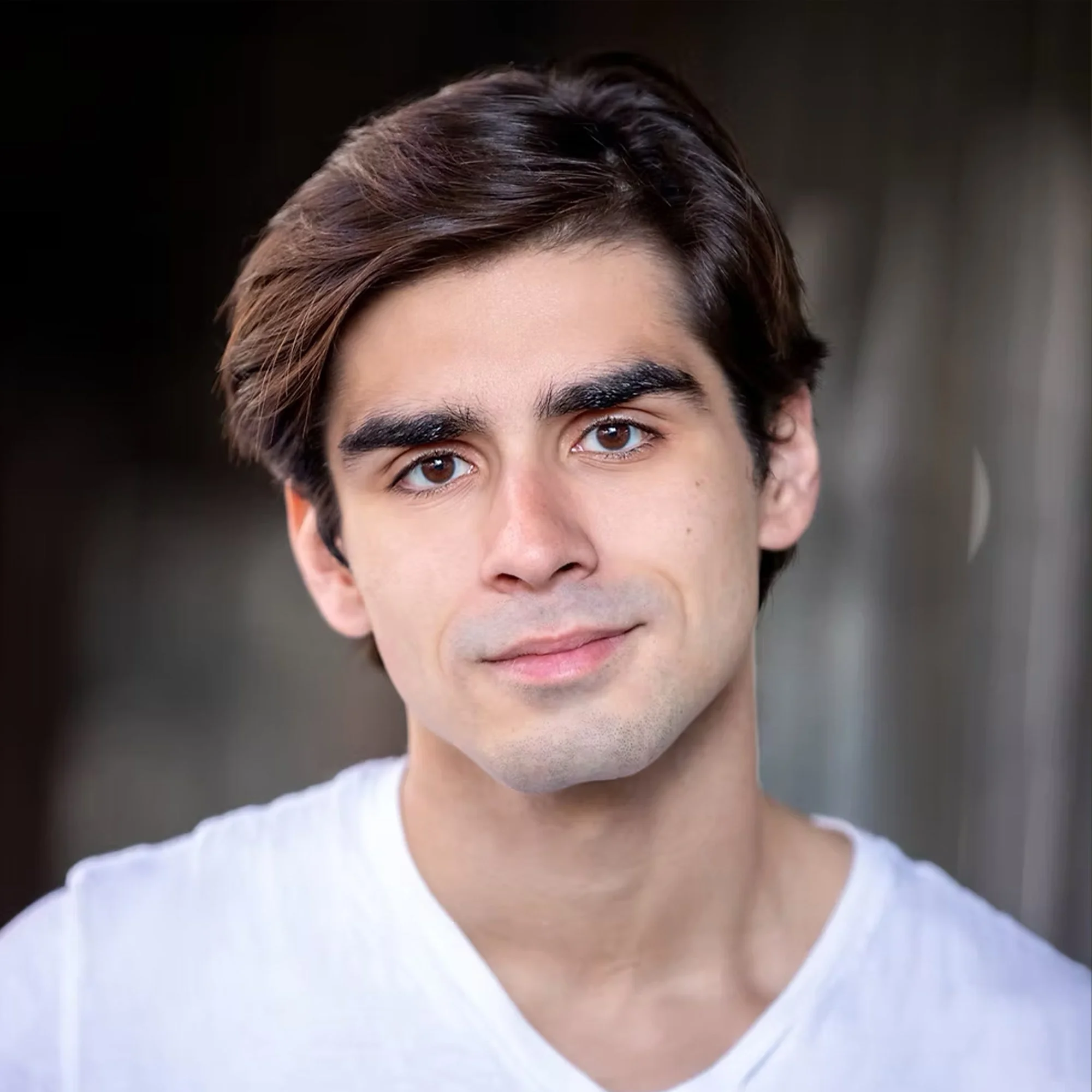 Close-up portrait of a young man with dark brown hair wearing a white shirt, smiling softly, against a blurred background.
