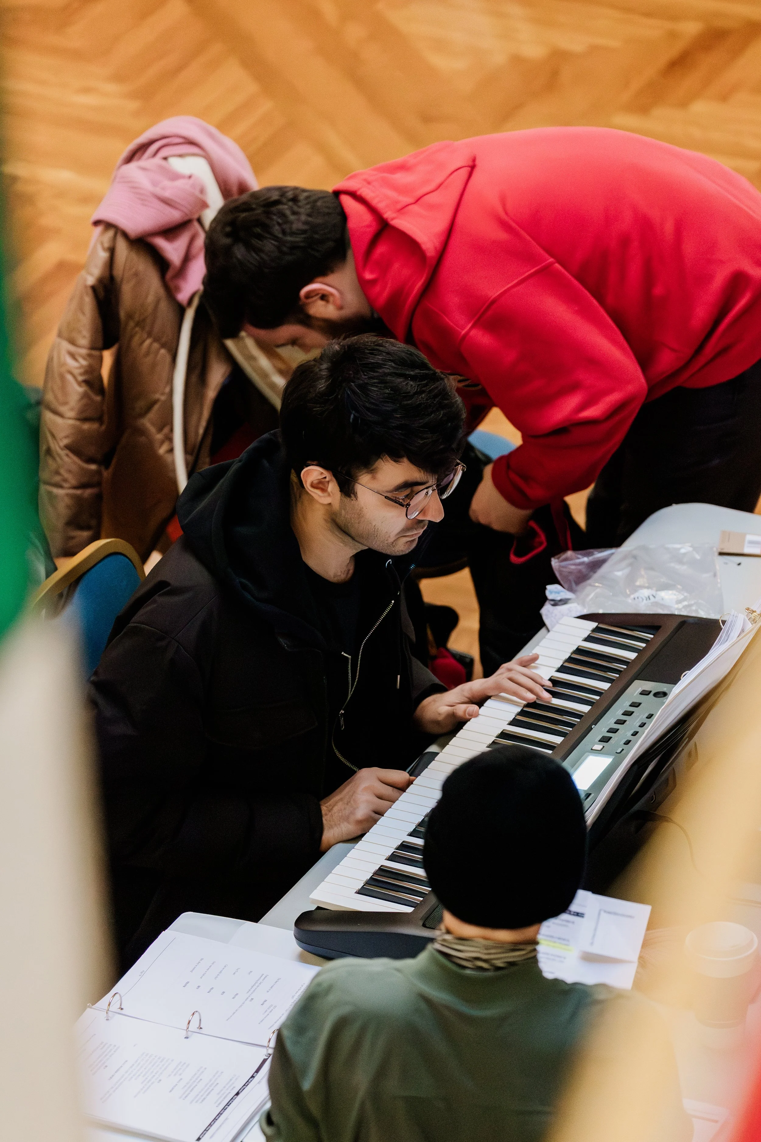 A person with glasses playing a keyboard or digital piano, with two individuals leaning over and observing, in a room with a wooden floor and table with papers and documents.