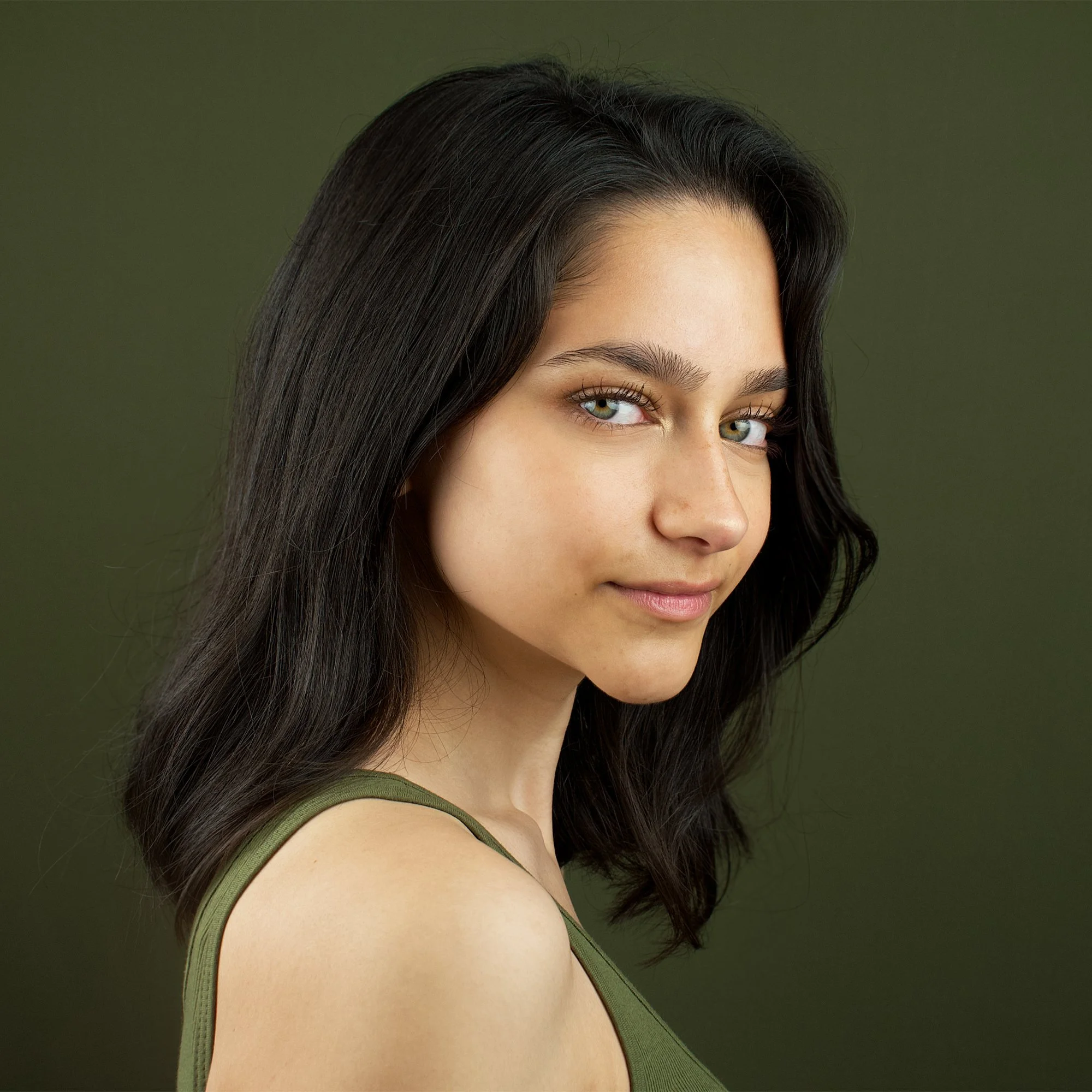 Close-up of a young woman with long dark hair, green eyes, and a slight smile, wearing a green tank top against a green background.