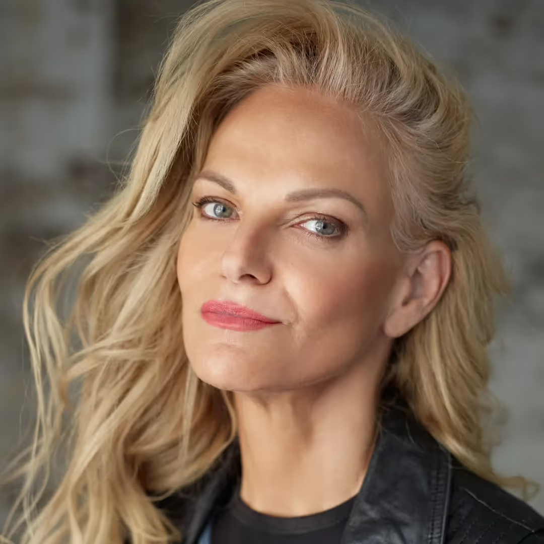 Close-up portrait of a middle-aged woman with blonde hair, blue eyes, and pink lipstick looking slightly to the side.
