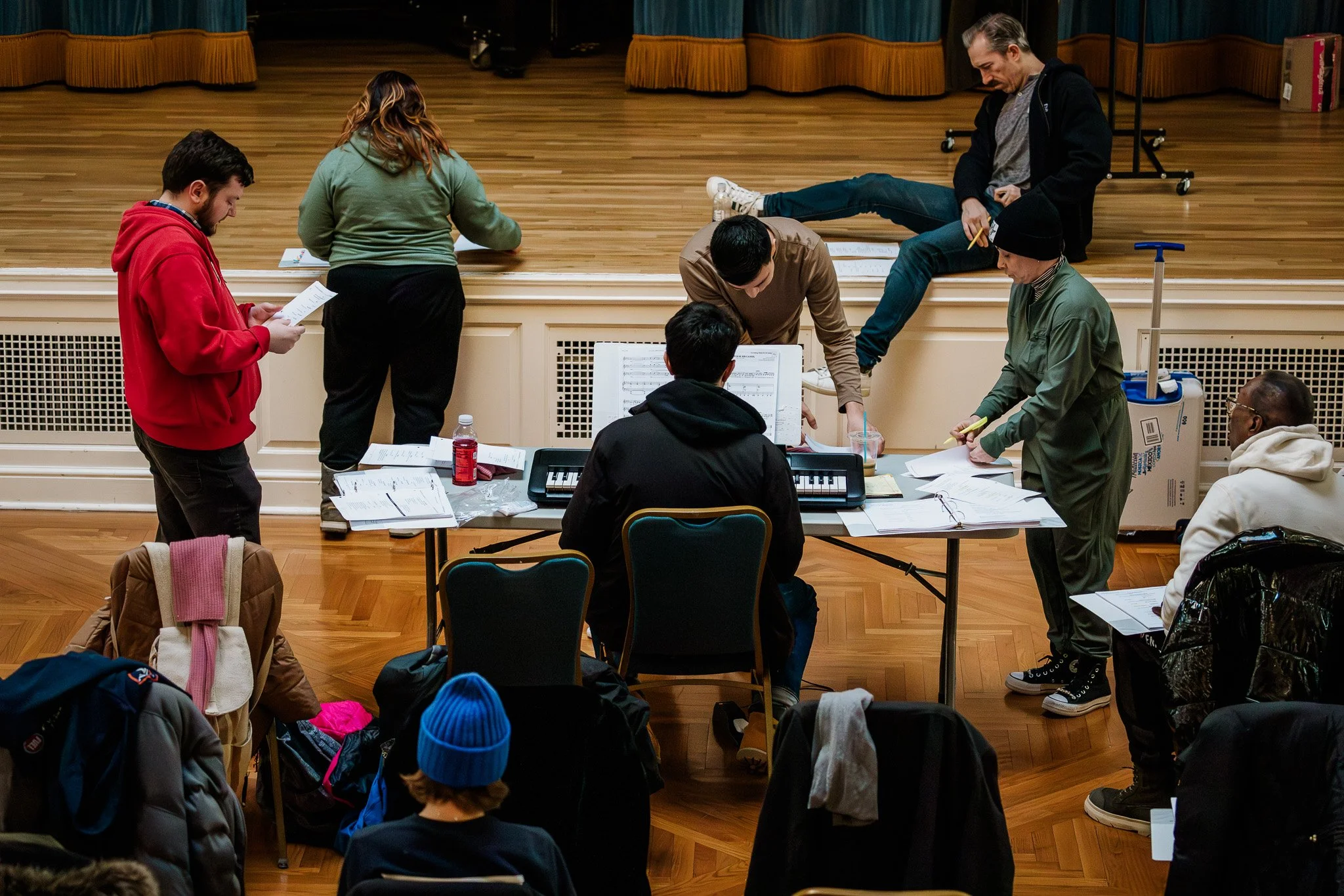 People practicing for a play or performance in an auditorium, with some sitting on stage and others working at a table with sheets of paper and music.