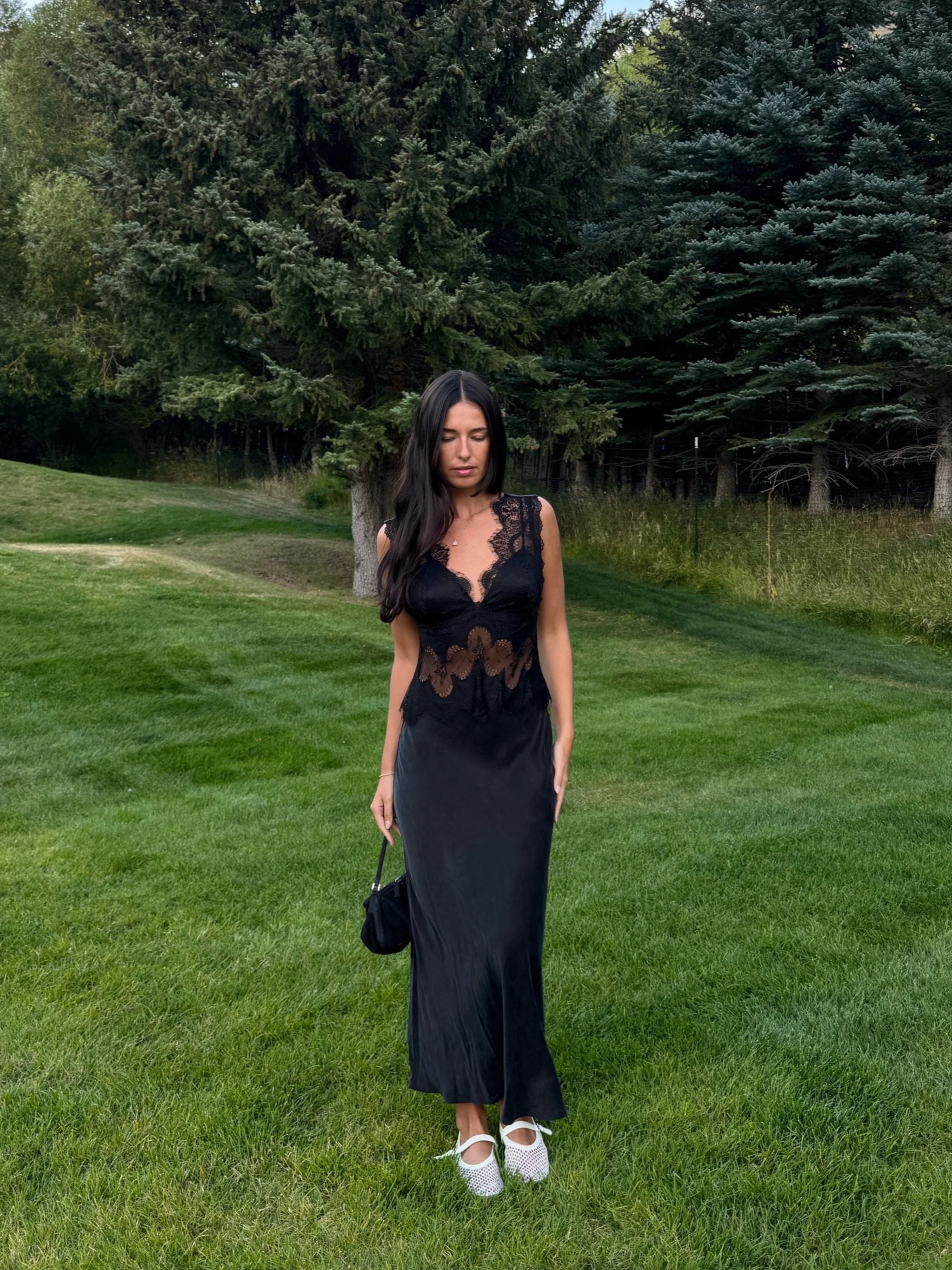 A woman in a black lace dress standing on grass in front of pine trees, holding a small black purse, and wearing white shoes.