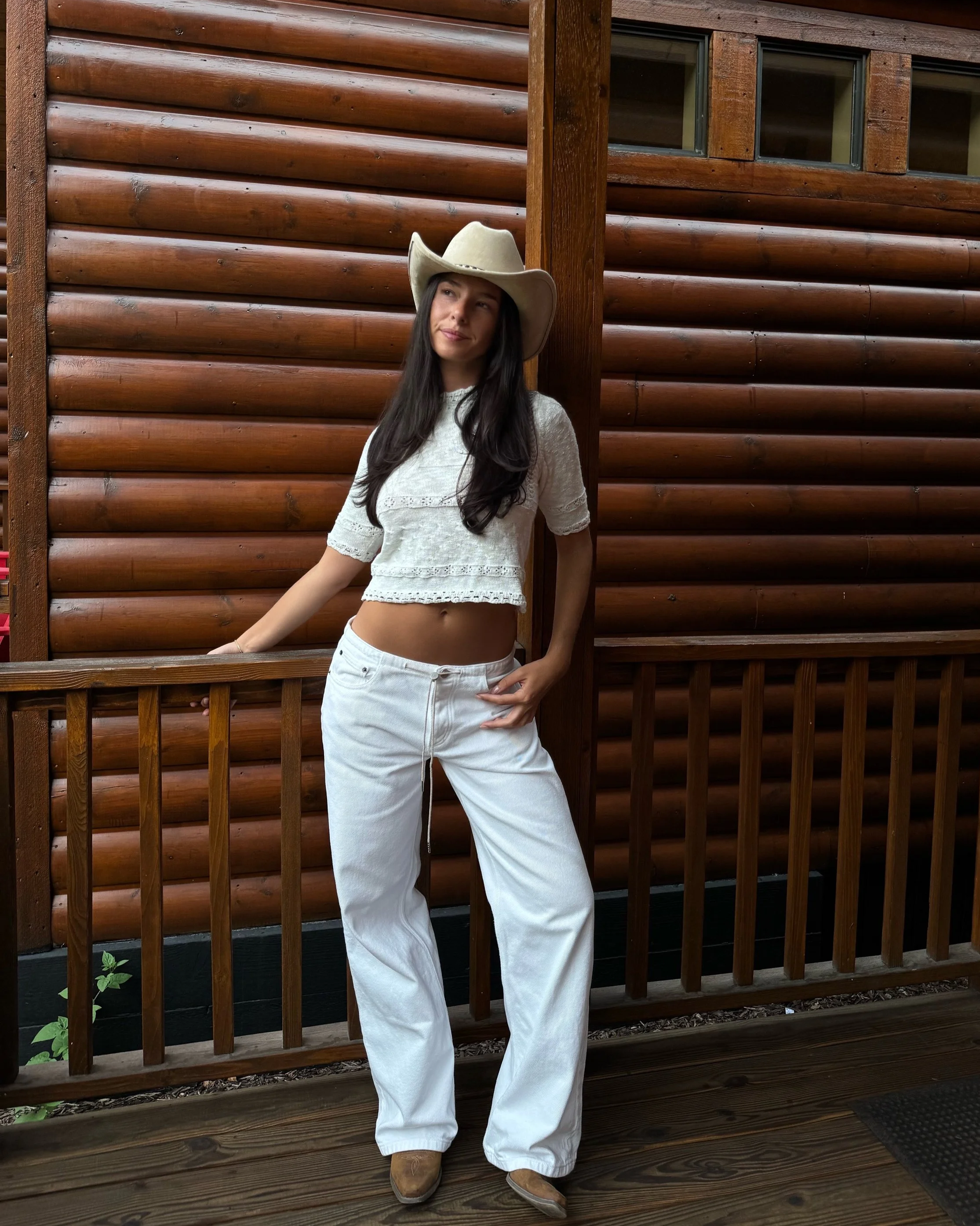 A woman wearing a cowboy hat, white crop top, and white jeans stands on a wooden porch with a log cabin background.