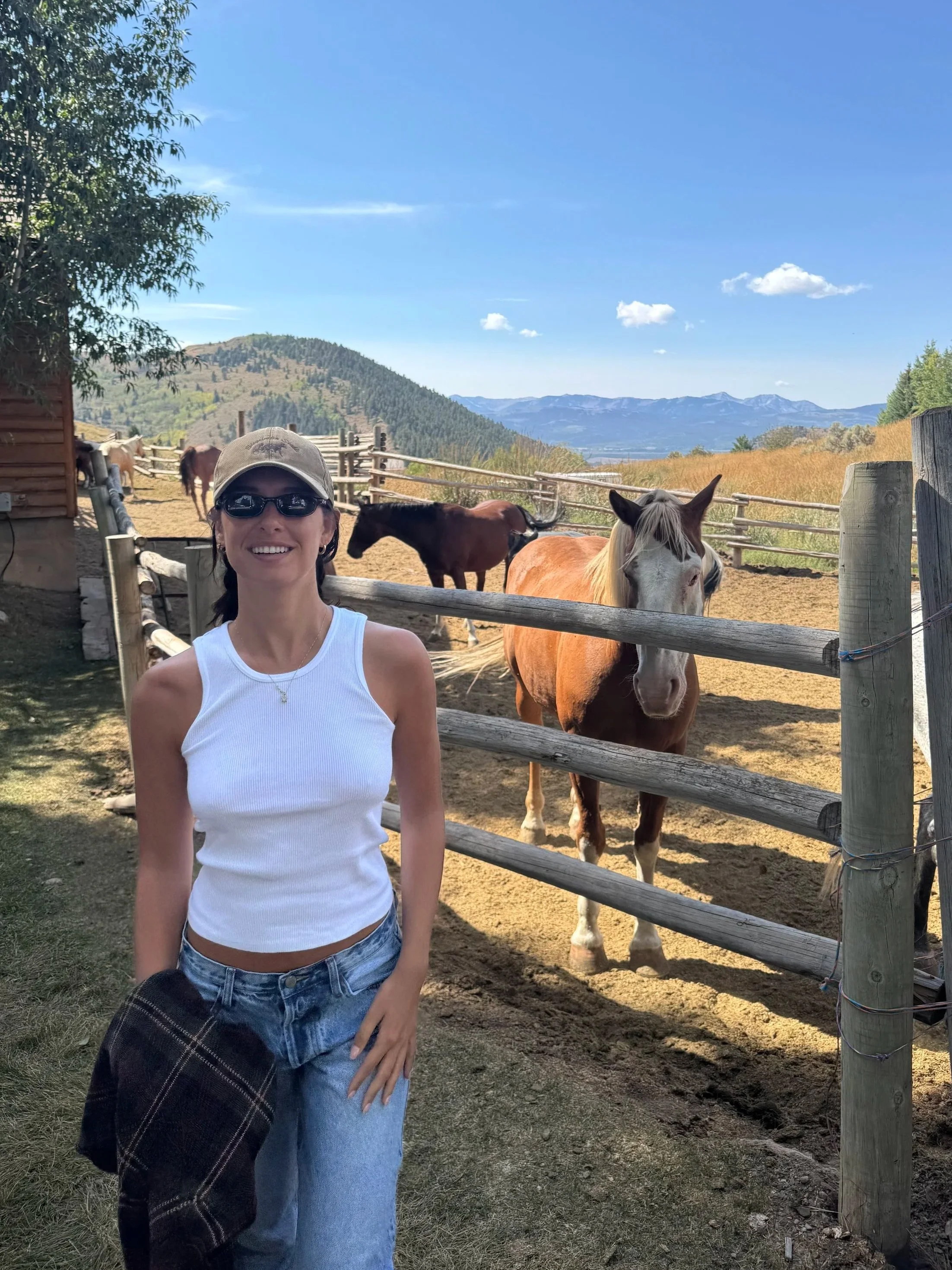 A woman standing outdoors in front of a wooden fence with horses behind her. She is wearing sunglasses, a beige cap, a white sleeveless top, and blue jeans, holding a black and brown plaid shirt. There are mountains and a partly cloudy blue sky in the background.