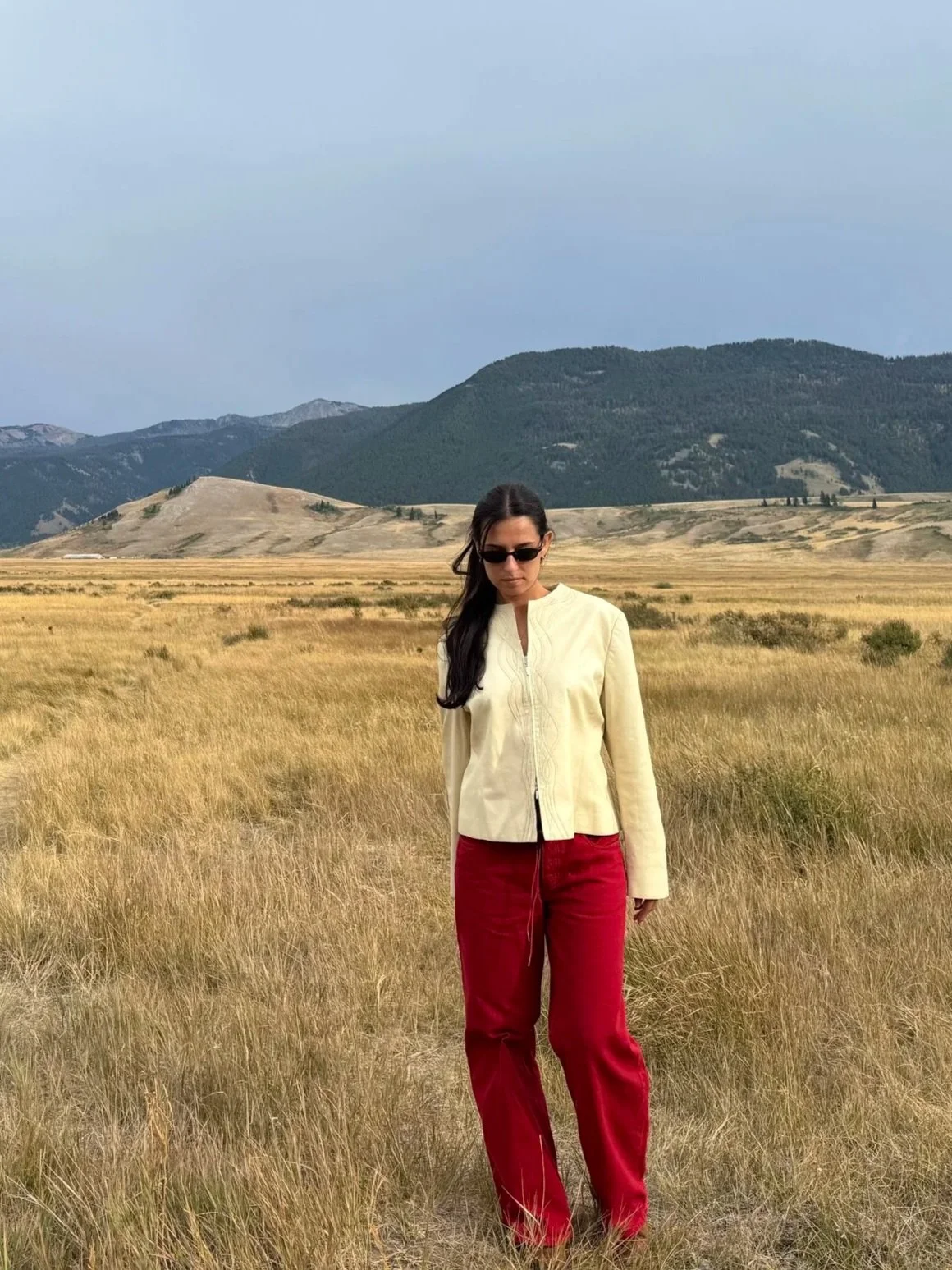 A woman with long dark hair wearing sunglasses, a cream-colored jacket, and red pants standing in a vast grassy field with mountains in the background under a cloudy sky.