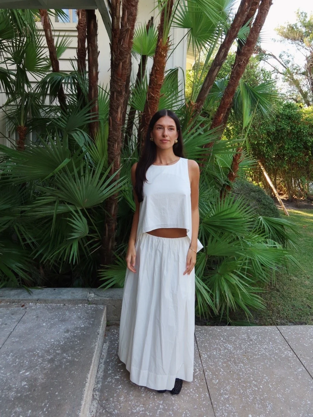 A woman in a white sleeveless crop top and matching long skirt standing outdoors in front of lush green tropical plants and trees.