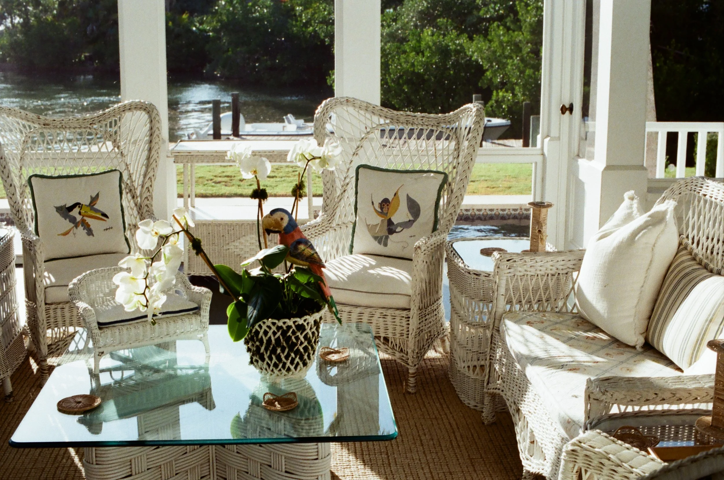 Sunlit screened porch with wicker furniture, including chairs and a loveseat, decorated with bird-themed cushions. A glass-topped table holds a vase of white orchids and a decorative bird figurine. Outside, a river is visible through the windows.